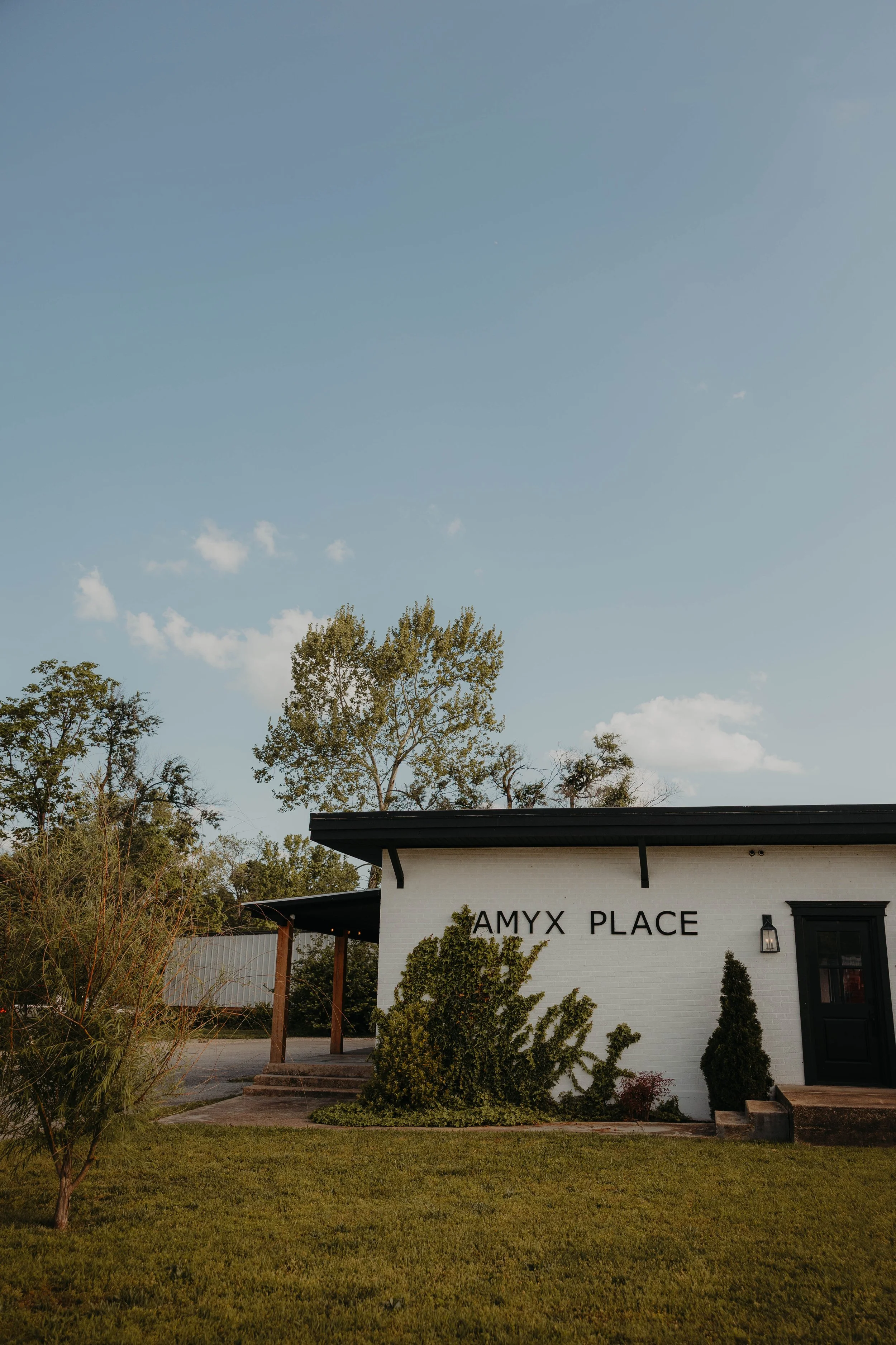 Exterior of a white building with black trim and the sign 'AMYX PLACE' on it, surrounded by green shrubbery and trees, with a blue sky overhead.