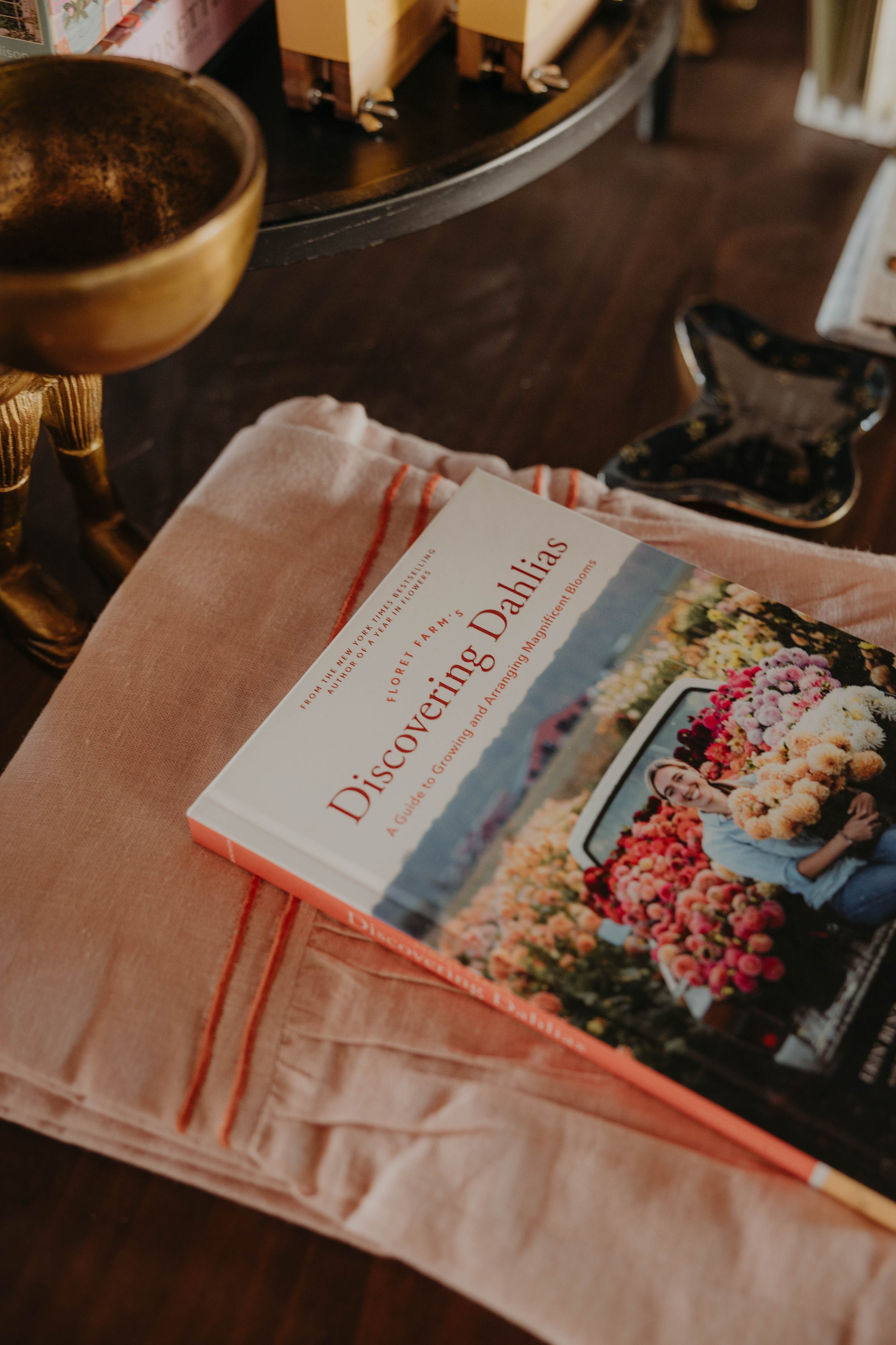 A book titled "Discovering Dahlias: A Guide to Growing and Arranging Magnificent Blooms" resting on a pink cloth. The cloth is placed on a table, with a brass bowl, wooden and metal furniture visible in the background.
