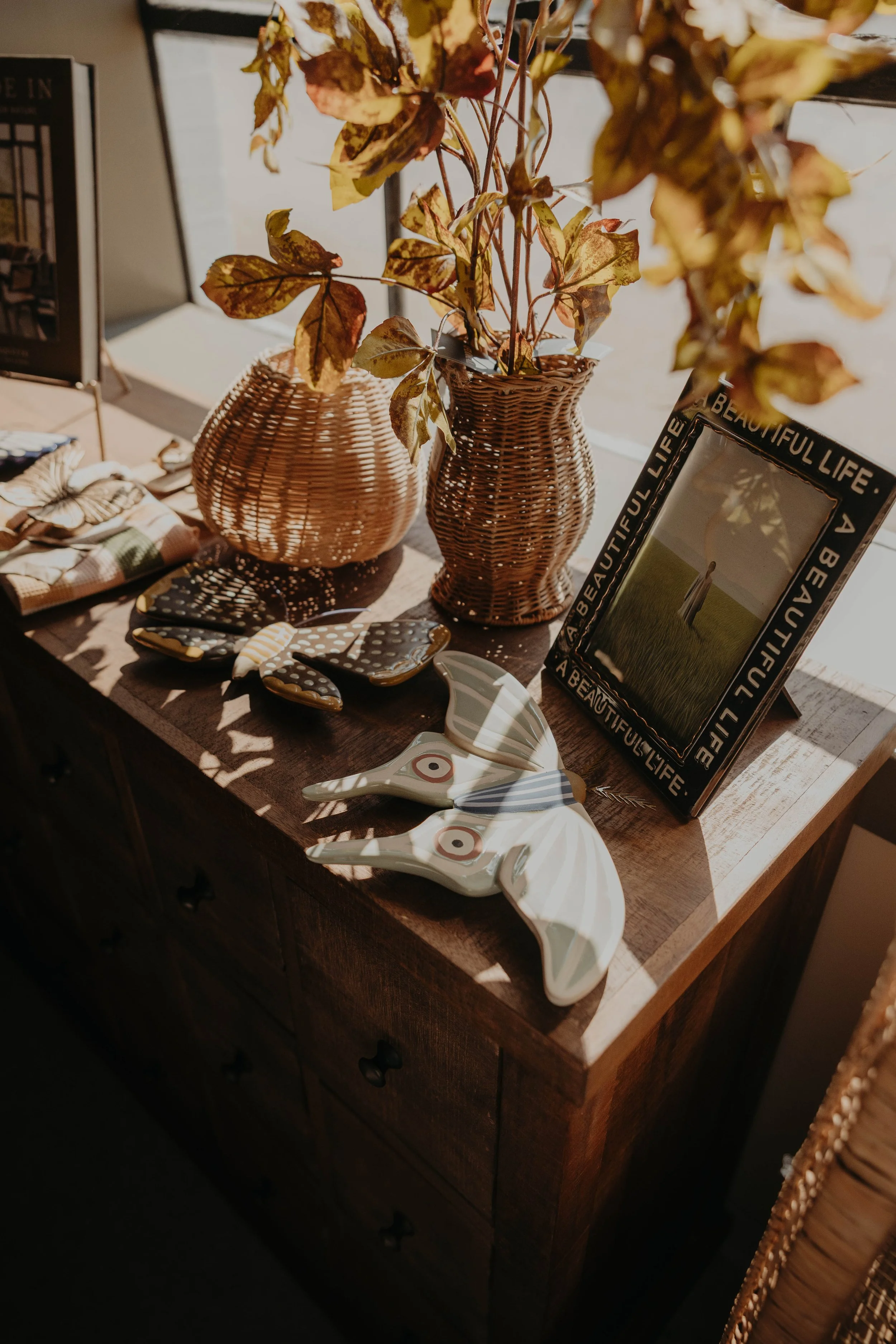 Decorative table with wicker vases, ceramic butterfly and bird figurines, framed photo with the words "Beautiful life" and accessories such as plates and napkins, sunlight casting shadows.