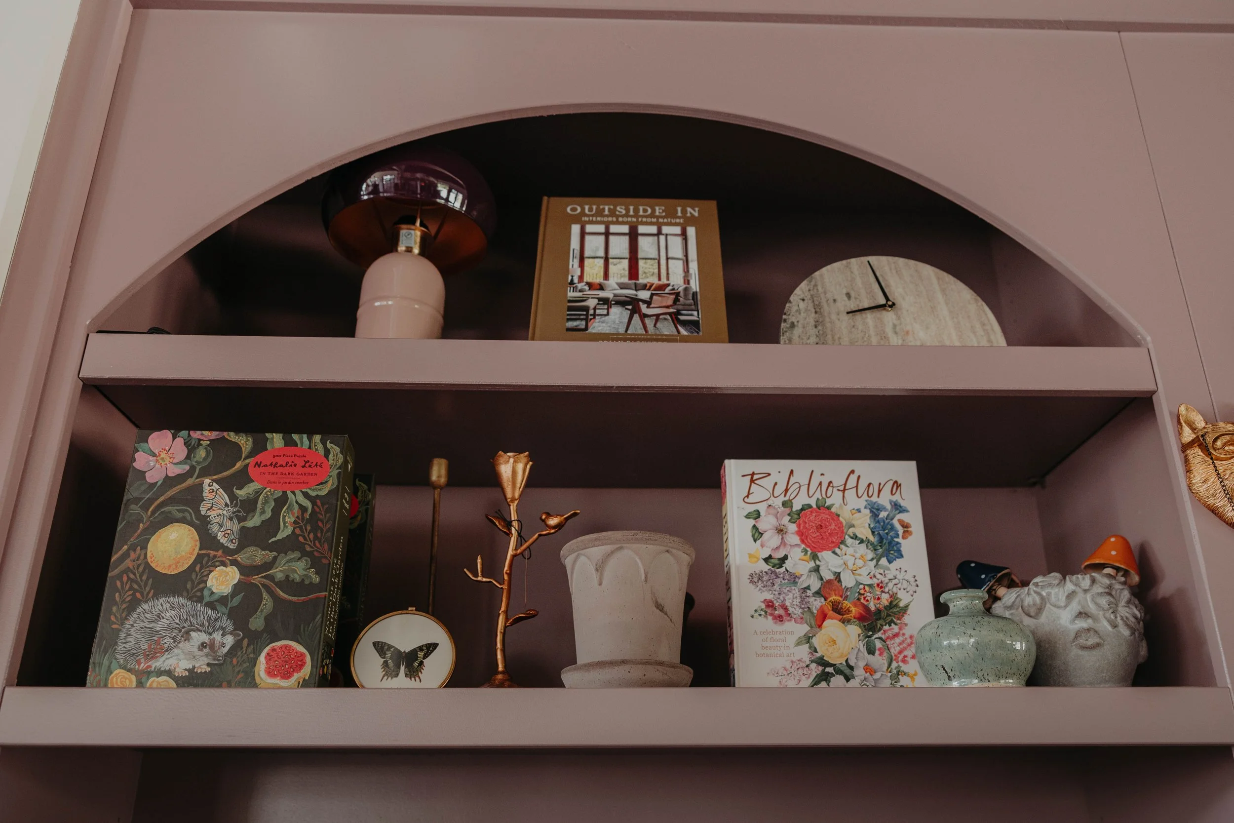 A pink shelf with a round pink lamp, a framed picture titled 'Outside In,' a white vase, a decorative gold tree, a butterfly coaster, a floral book titled 'Biblioflora,' a ceramic container, and a small orange item.