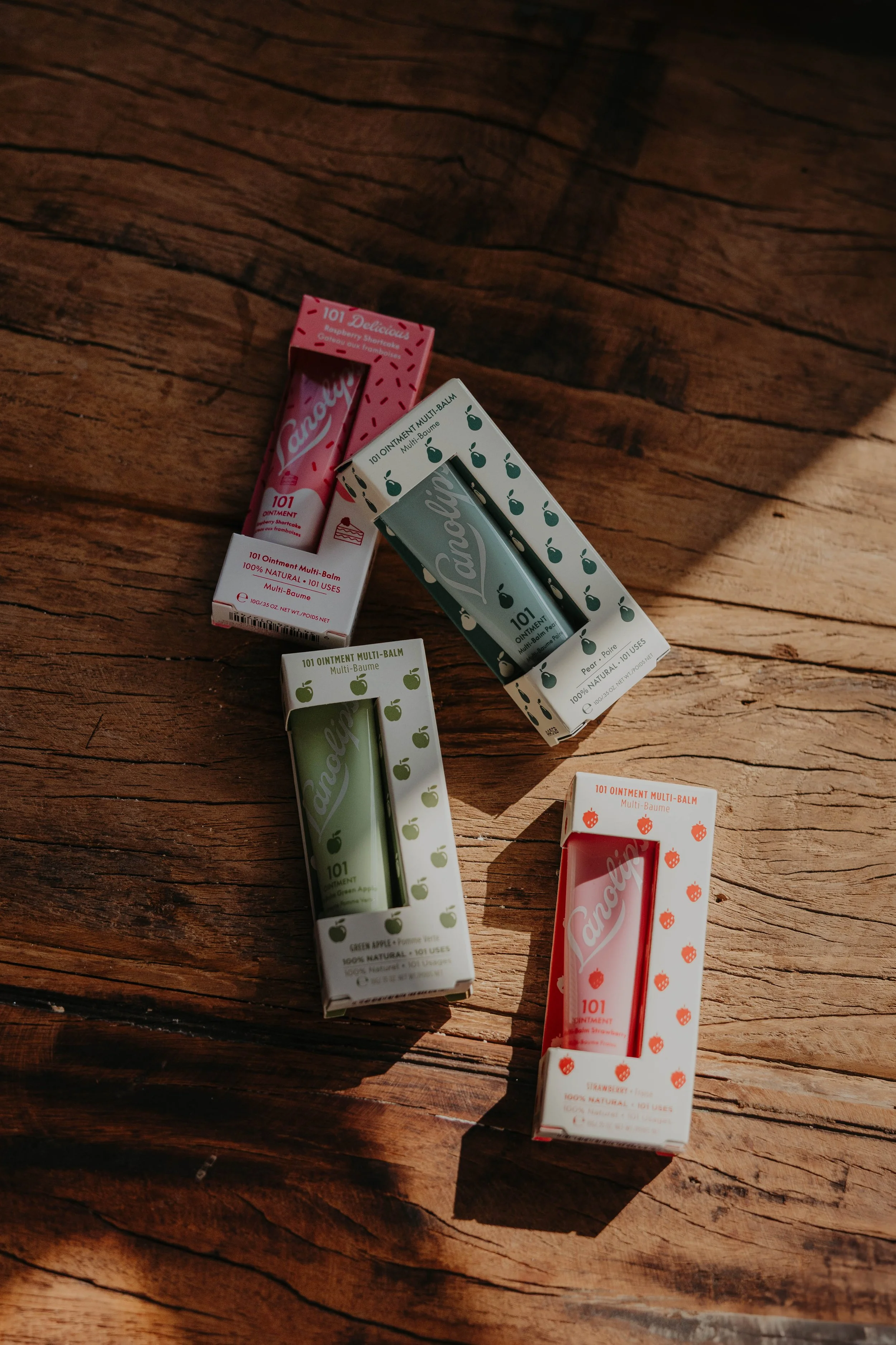 Several colored lip balm tubes and their boxes on a wooden surface, featuring flavors like strawberry, green apple, and raspberry.