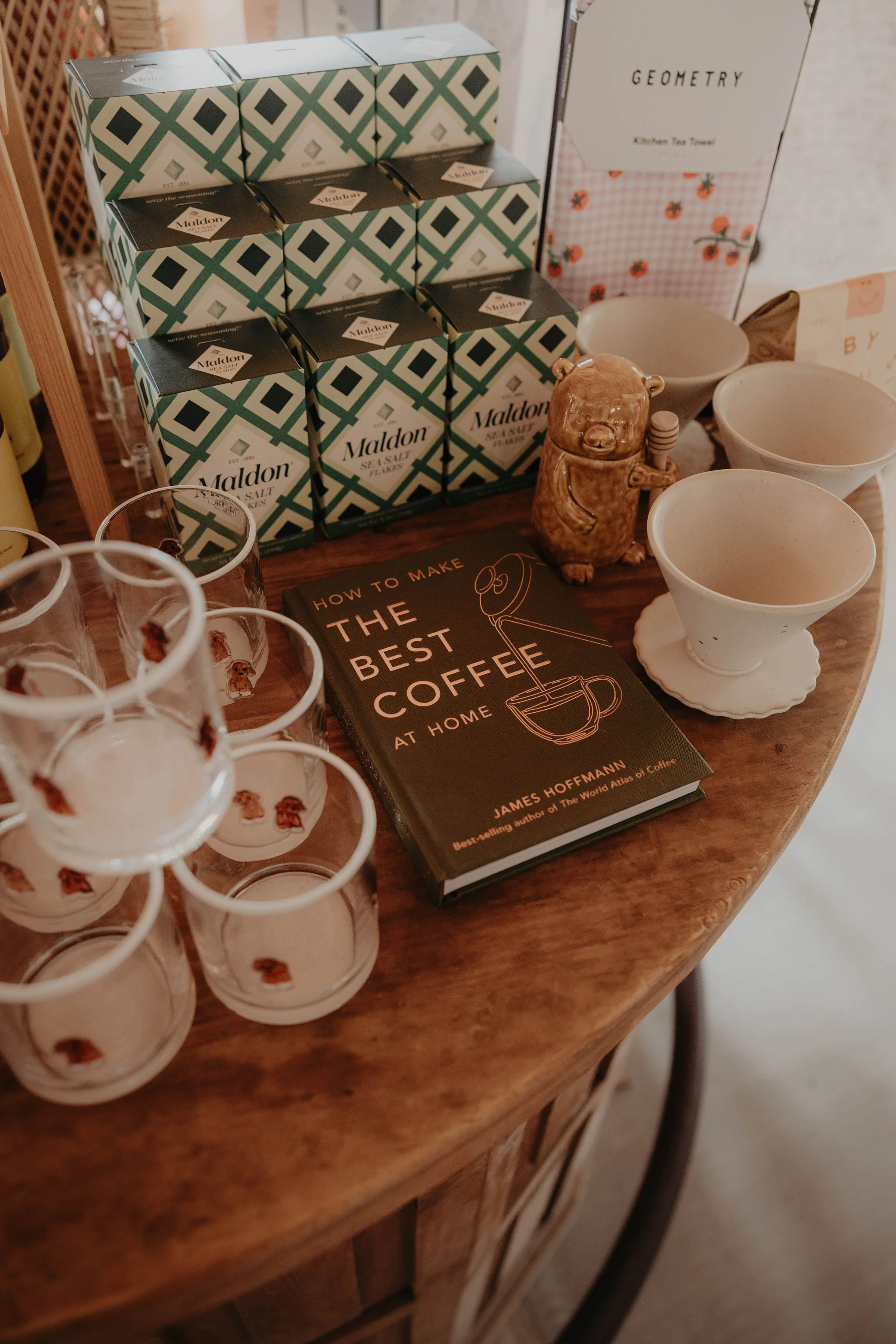 Stacked boxes of Maldon sea salt, a book titled "How to Make the Best Coffee at Home" by James Hoffmann, three white bowls, a bear-shaped ceramic figure, and a glass with a cartoon dog print, all on a wooden table.