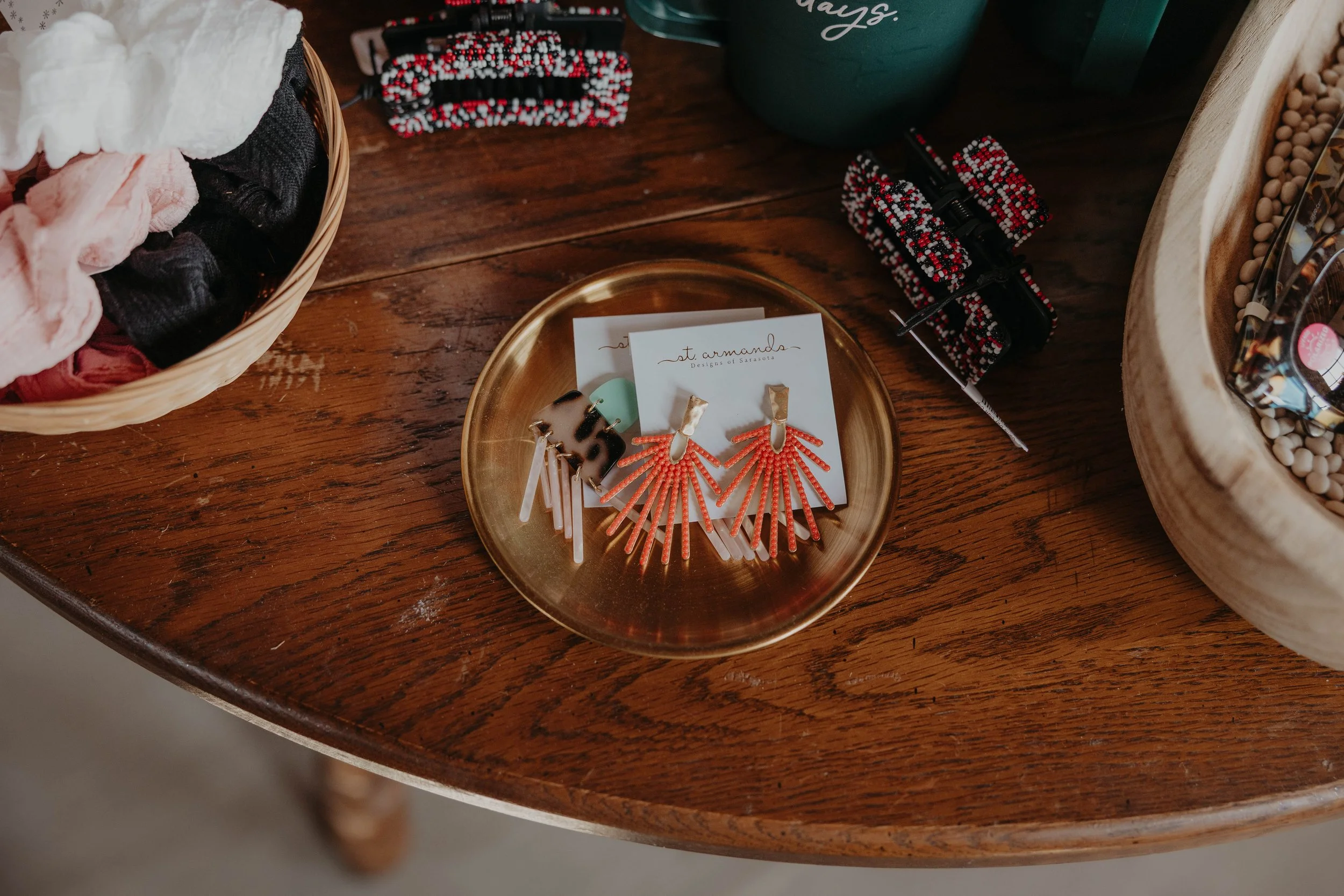 Decorative earrings on a white card, placed on a gold tray on a wooden table, with a basket of cloth items, and other accessories nearby.