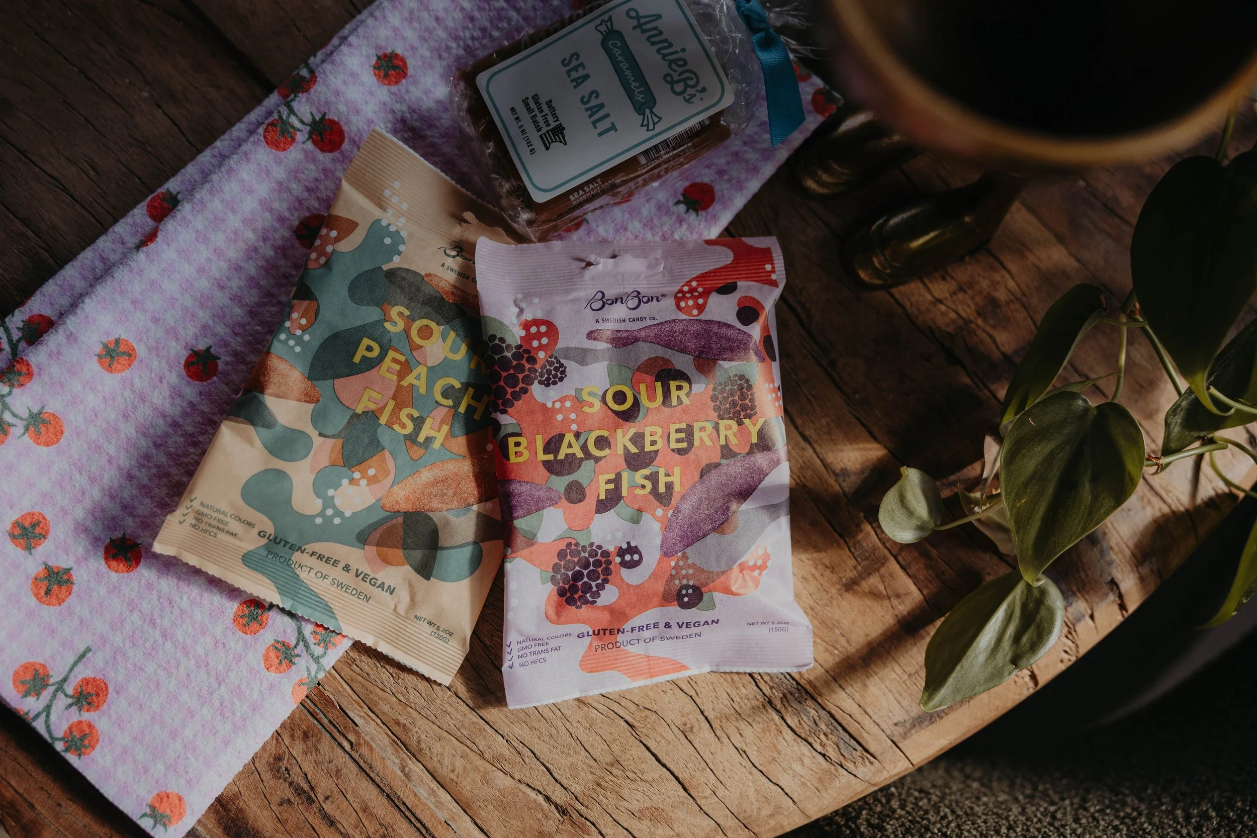 A wooden surface holding a pink towel with tomato prints, a packet of sour peach fish candy, a packet of sour blackberry fish candy, a jar of sea salt, and some green leaves.