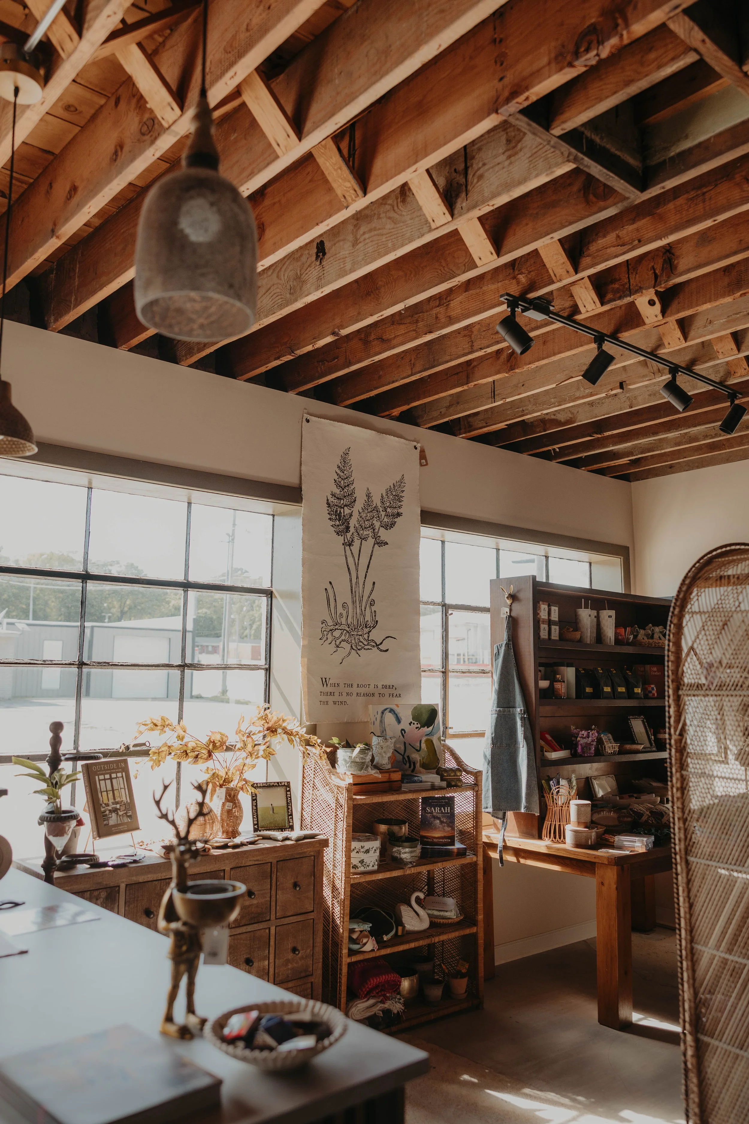 Interior of a cozy shop with wooden shelving and decor, large windows letting in natural light, hanging lamps, and various plants and books displayed.