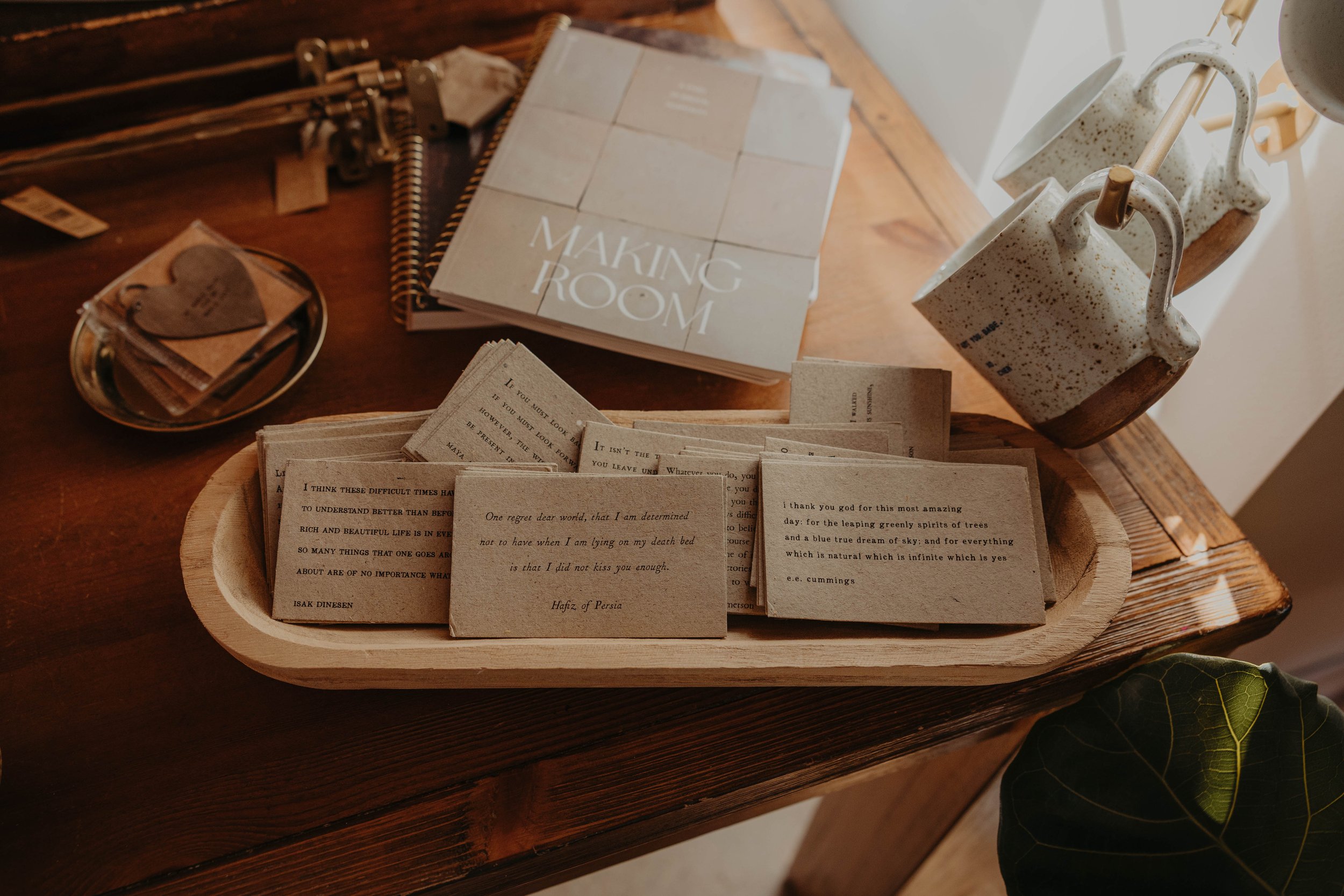 A wooden table displaying a cardboard box filled with small paper cards, a spiral-bound notebook titled 'Making Room,' a small dish with tags, and decorative speckled mugs hanging on hooks.