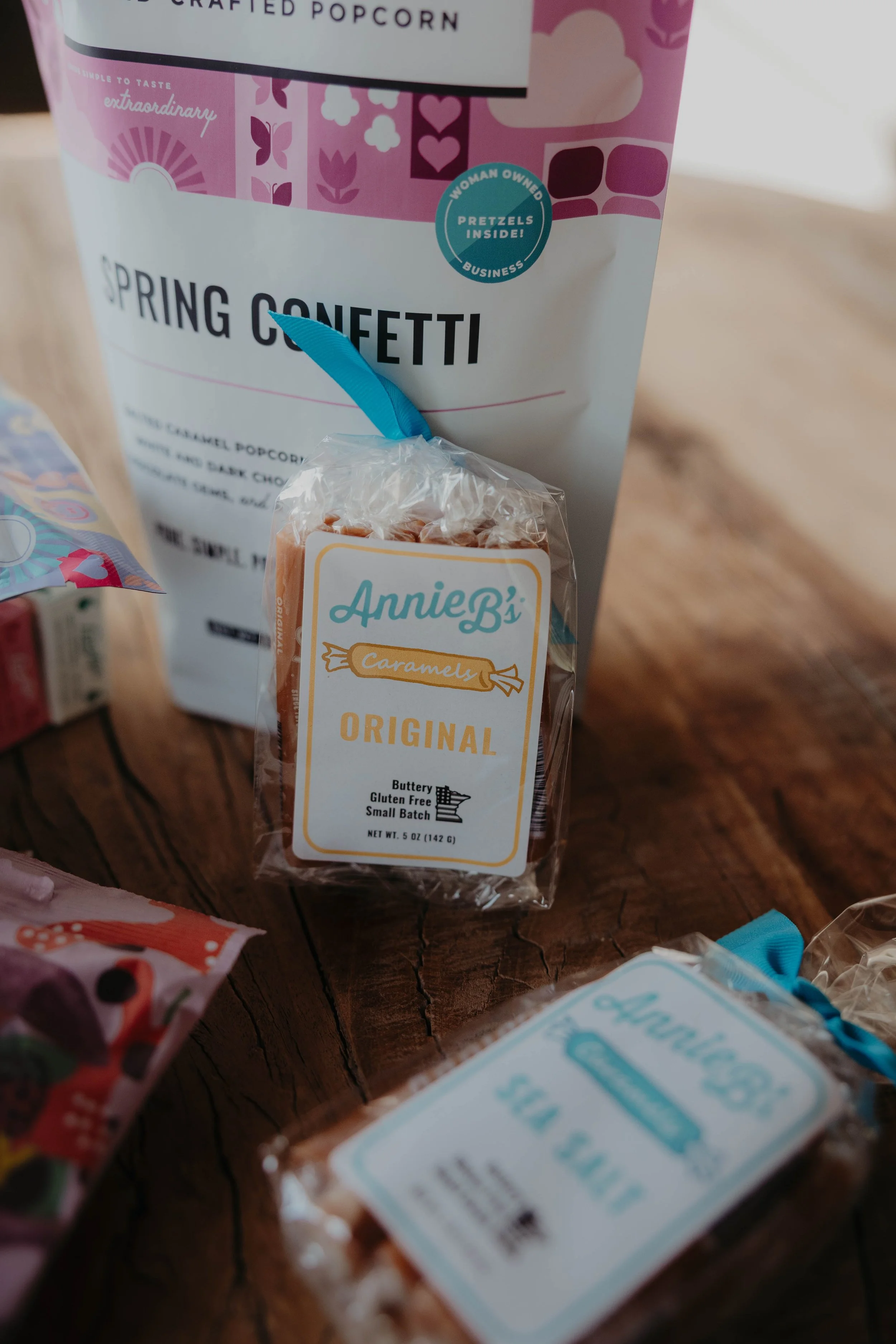 Close-up of snack items on a wooden surface, including a bag of Annie B's caramel candies and a bag of popcorn labeled 'Spring Confetti' with pink and purple packaging.
