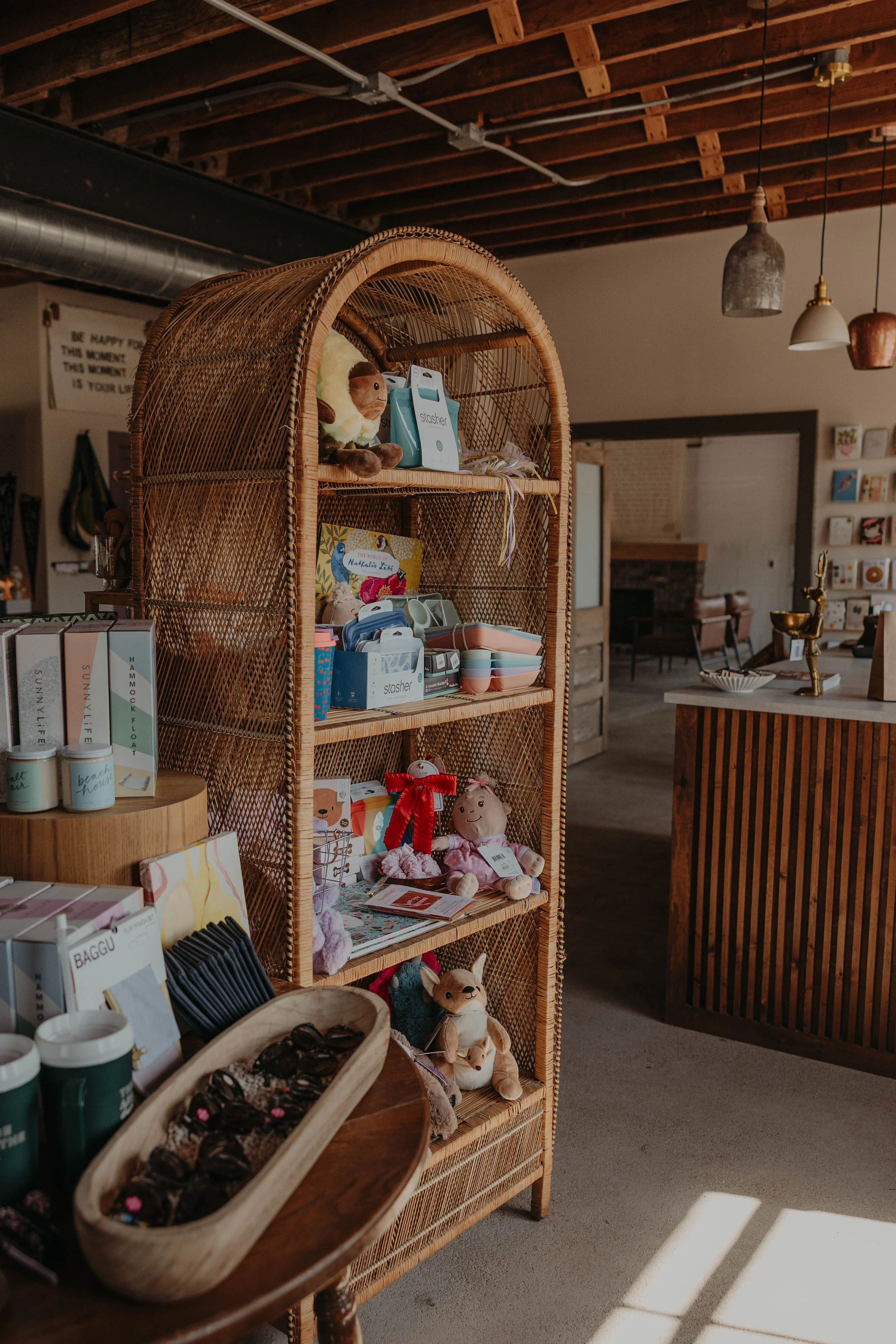 Interior view of a gift shop with a wicker display shelf filled with plush toys, books, and small accessories. The ceiling has exposed wooden beams and hanging pendant lights, with part of a table and other store items visible.