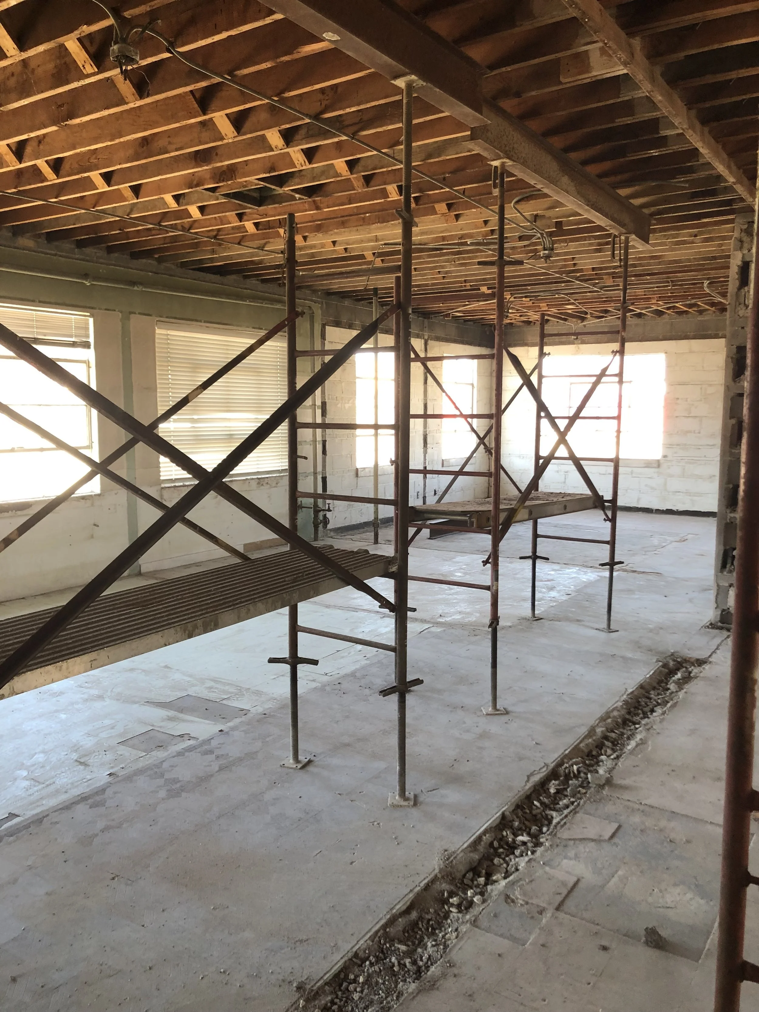 Interior view of a building under construction, with exposed ceiling beams, scaffolding, and unfinished flooring.