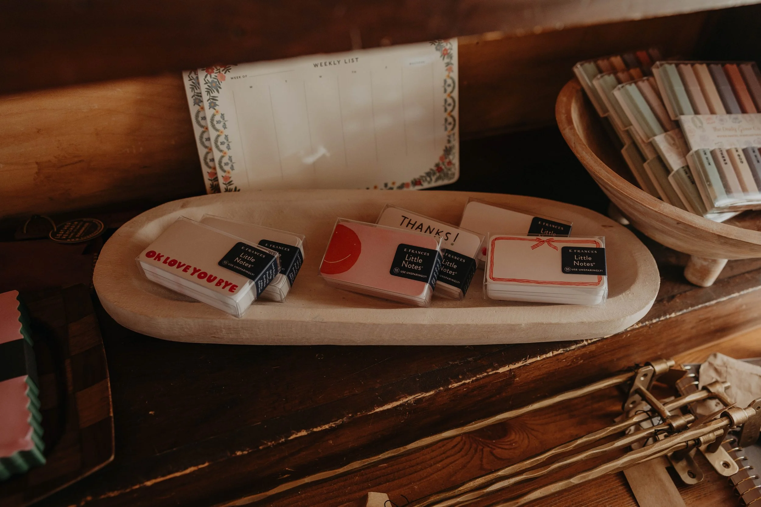 A wooden table displays small boxes of notecards, some with messages like 'THANKS!', 'OK LOVE YOUR YBE', and 'Little Notes', along with a weekly planner with a floral border partially visible behind the notecards.