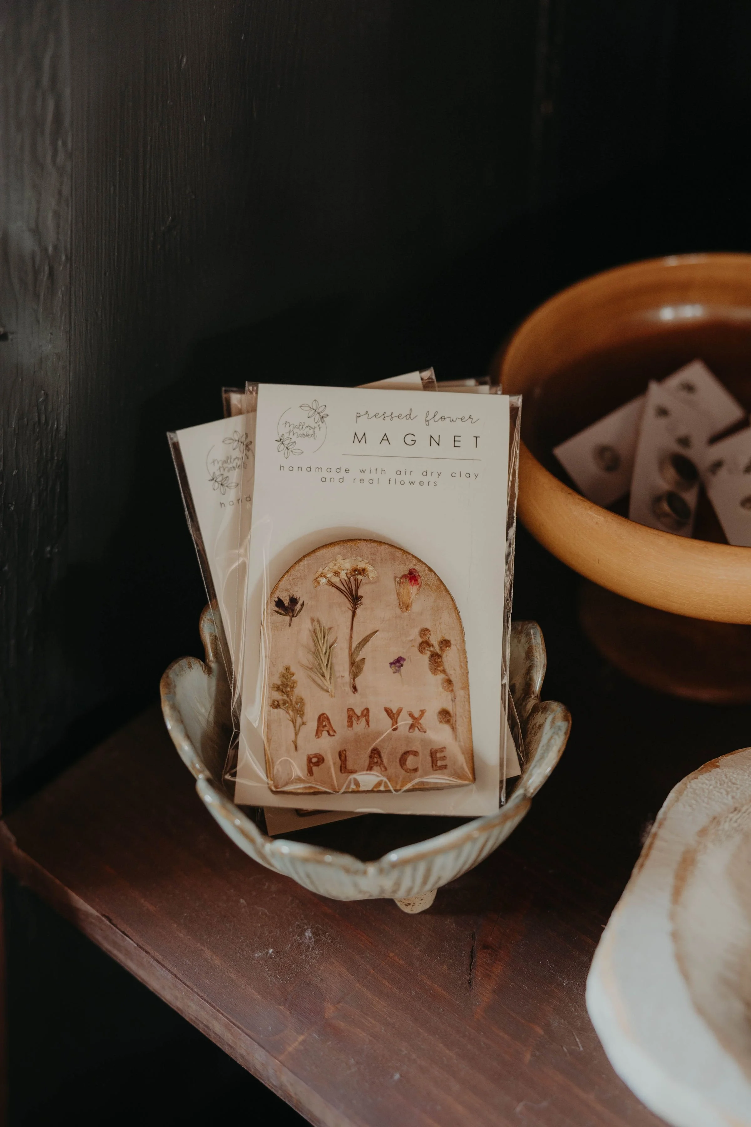 A small paper card with pressed flowers inside a basket on a wooden surface, with another card behind it that says "pressed flower MAGNET" and features illustrations of flowers.