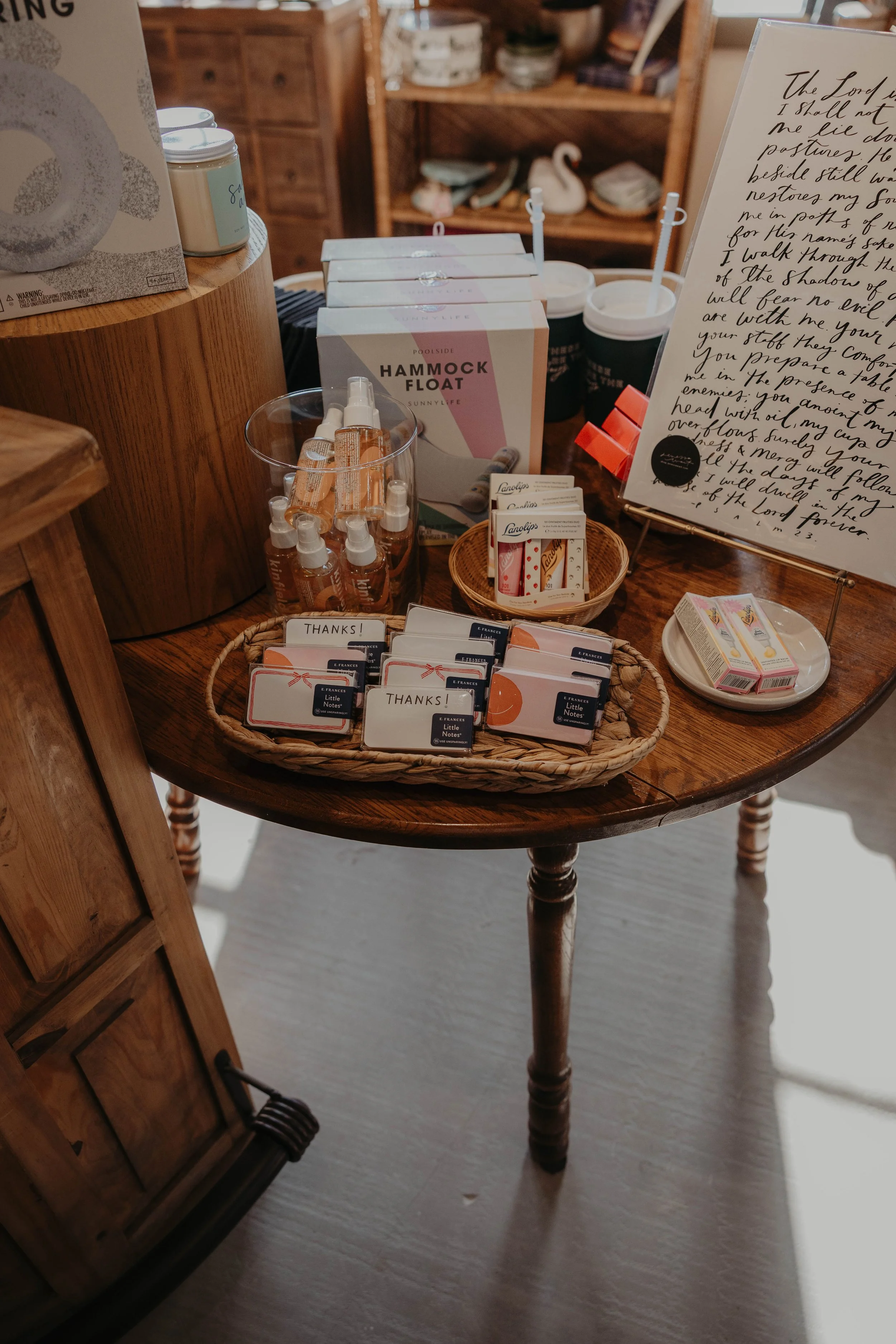 A round wooden table displaying various items including small bottles, thank you cards, packaged lip balms, a boxed hammock float, and a large decorative sign with handwritten text. In the background, shelves hold additional decorative objects and bo
