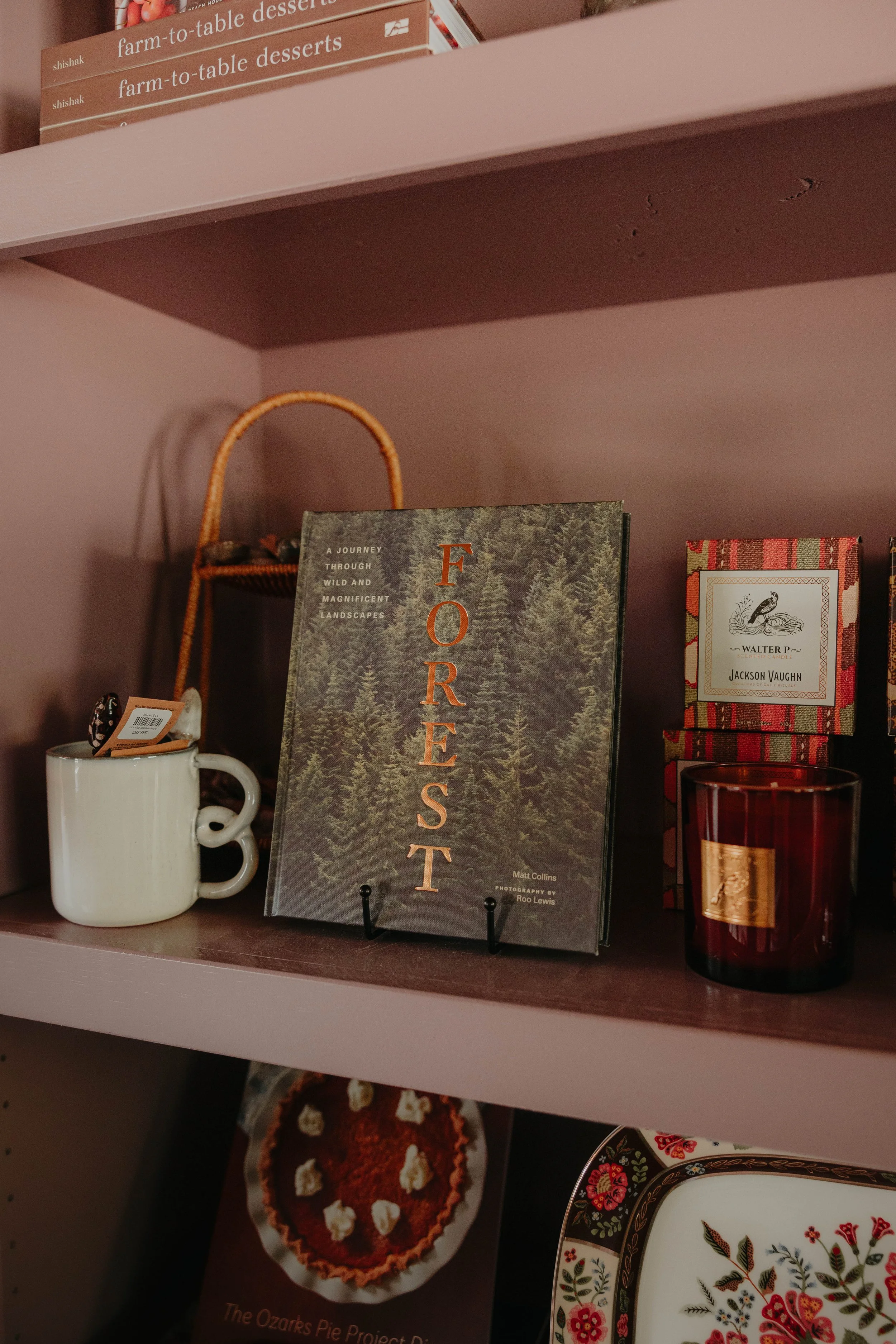 A pink bookshelf with books titled 'Farm-to-Table Desserts,' a decorative book about forests, a box of Jackson Vaughn candles, a white mug, and a red candle holder with a gold label.