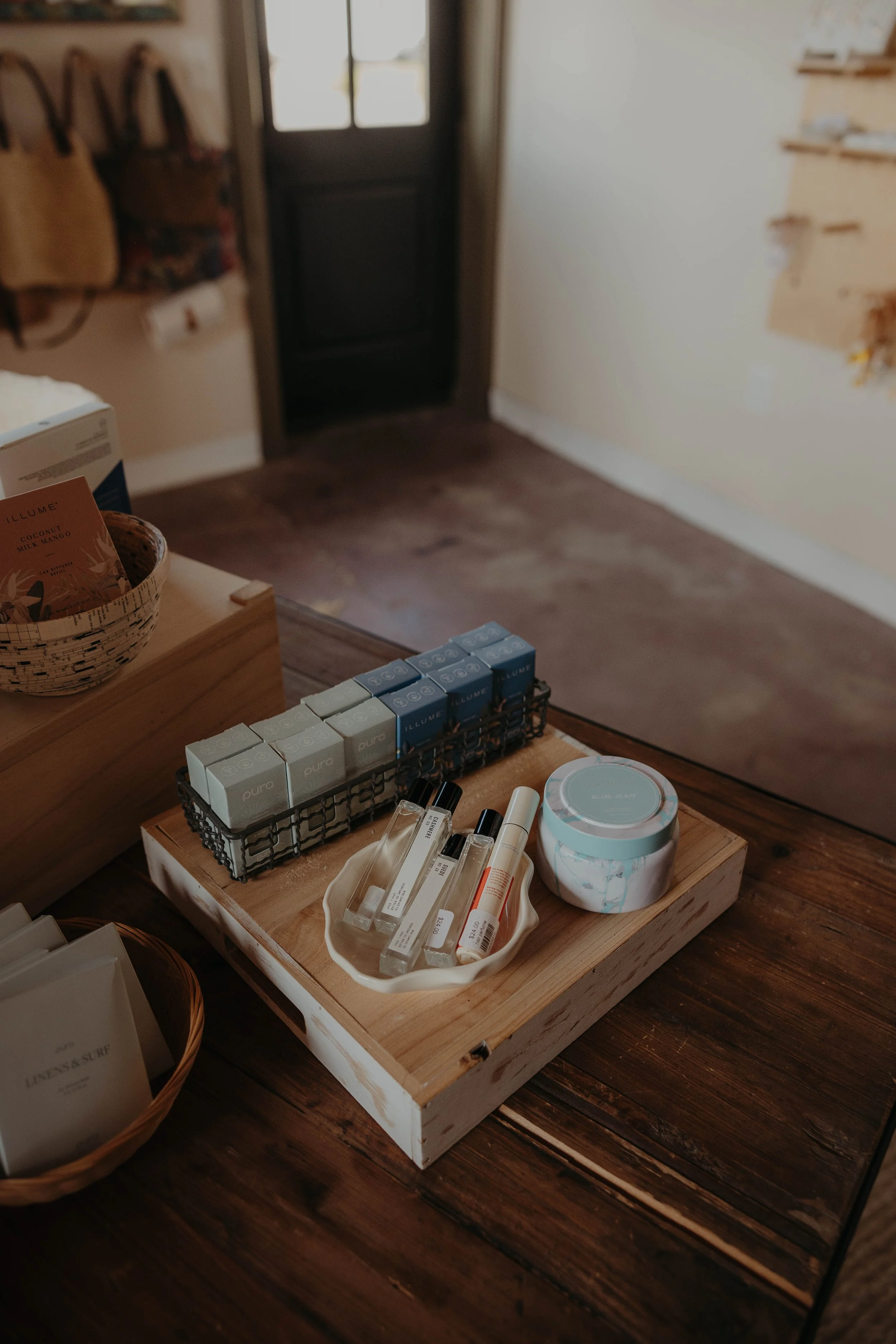A wooden display table with skincare products, including bottles, tubes, and boxes, on a wooden surface. In the background, a black door with glass panels is partially visible, with a wall on the right and some shelves on the left.