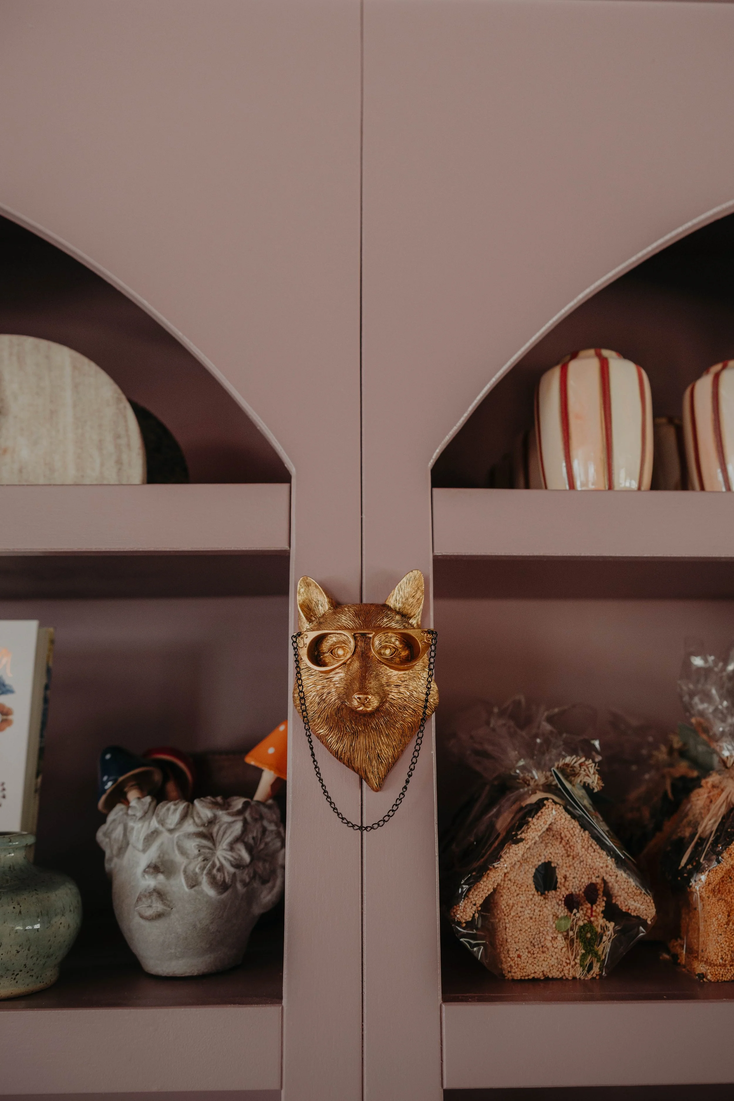Decorative metal cat face with glasses necklace hanging on a pink shelf, with vases and other decorative items in the background.