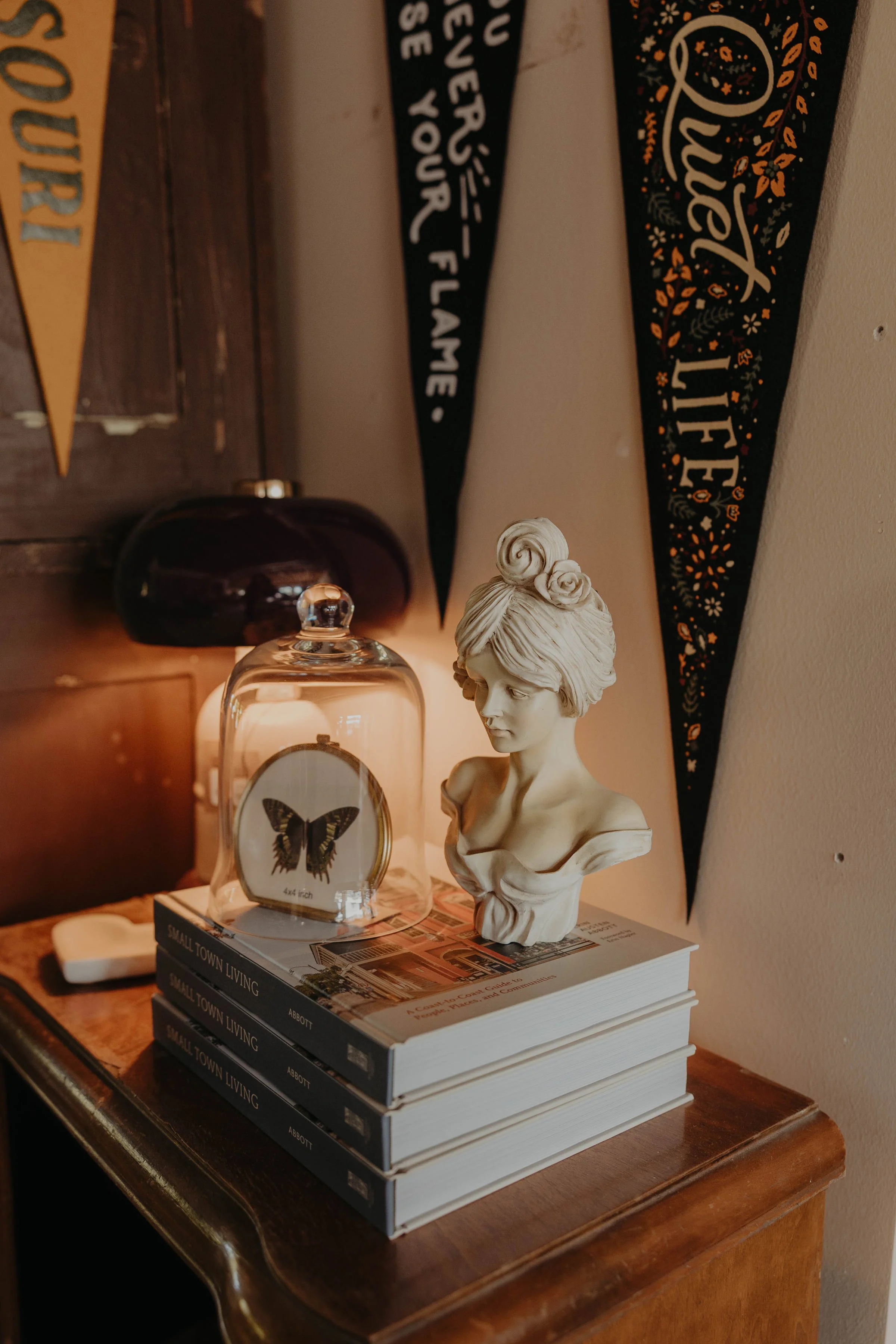 A decorative arrangement on a wooden side table featuring a stack of five books titled 'Small Town Living,' a glass display case with a butterfly illustration, a vintage-style bust sculpture of a woman, a black table lamp, and a dark decorative banner with floral patterns hanging on the wall.
