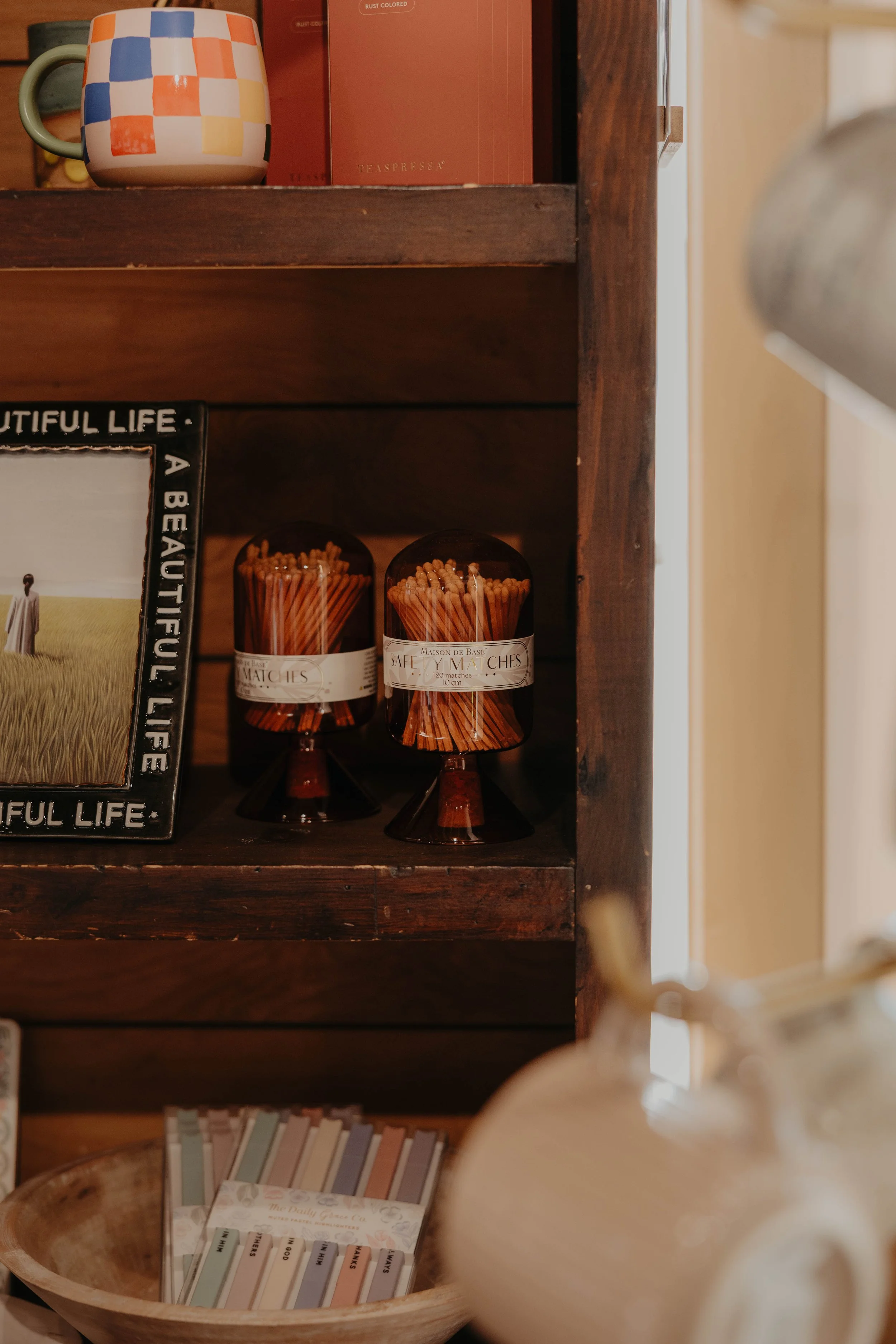 A wooden bookshelf containing various items including a mug with a colorful checkered pattern, red books, a metal photo frame with the words 'Beautiful Life' on the border, jars of matches, and a tray of assorted colored index cards.