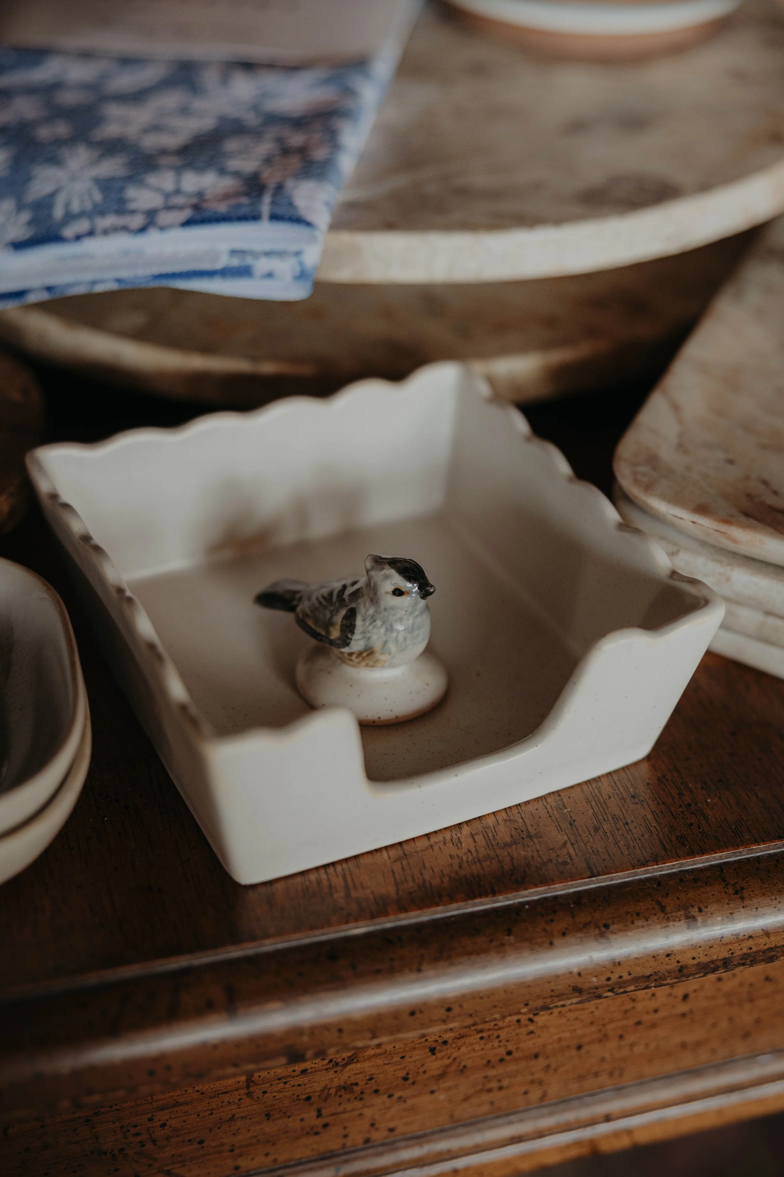 A small ceramic figurine of a bird with black feathers and black nose inside a square white ceramic dish with scalloped edges, placed on a wooden surface.