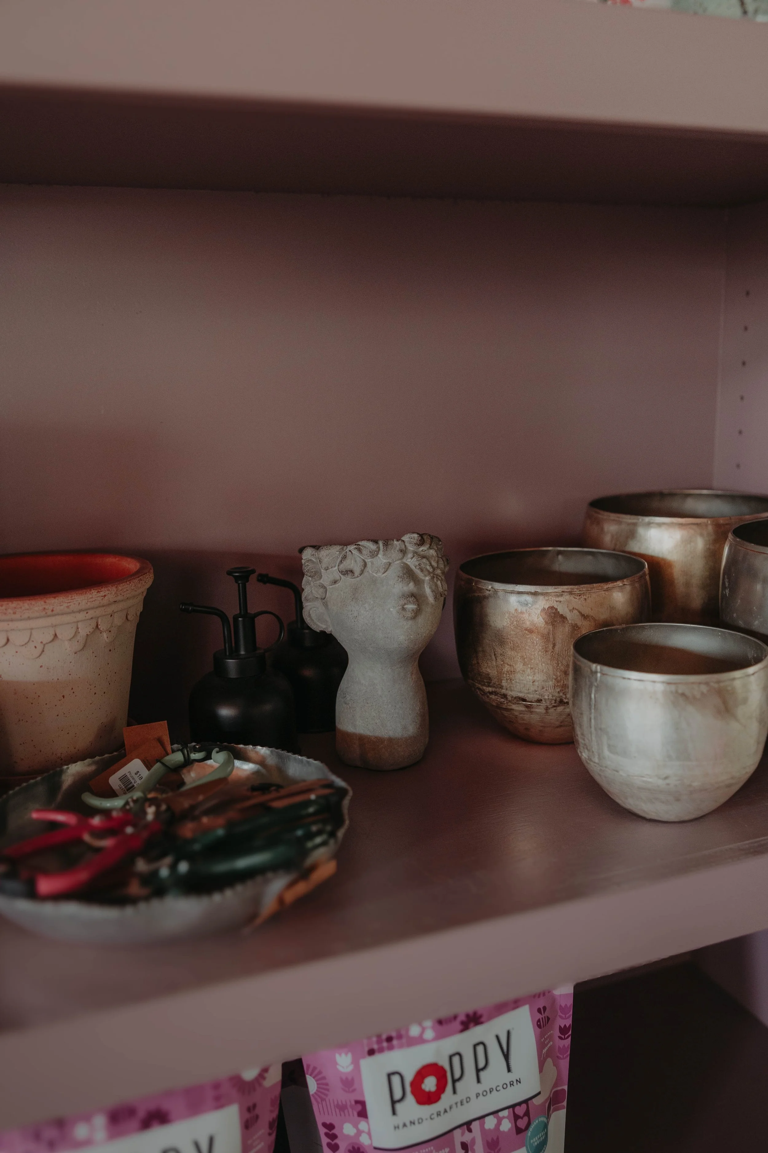 A shelf with various ceramic bowls and vases in neutral colors, a black soap dispenser, and a decorative porcelain piece. Below the shelf, there are pink popcorn bags labeled 'Poppy Hand-Crafted Popcorn'.