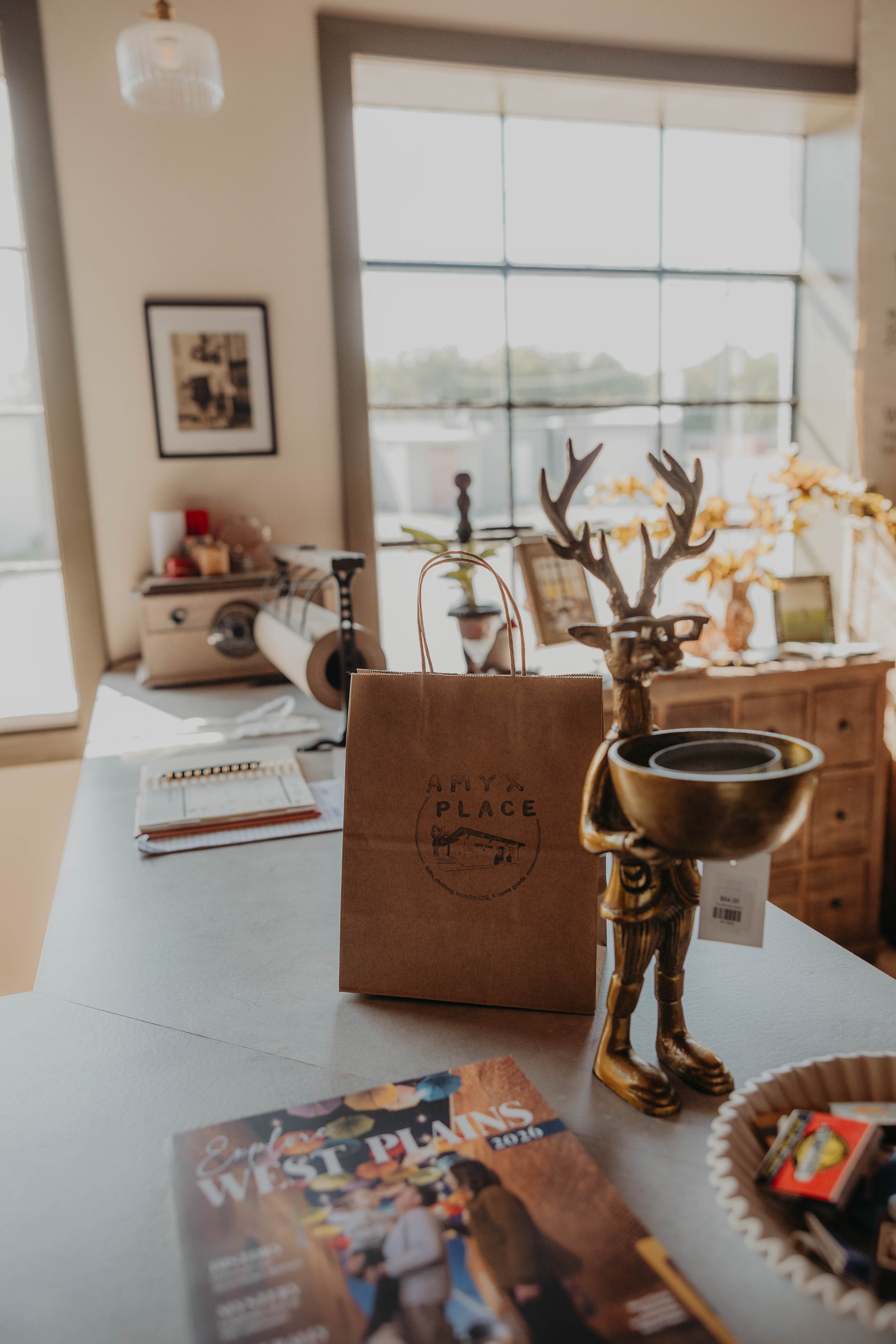 Indoor scene with a desk displaying a paper bag with the logo 'Amy's Place,' a decorative reindeer with large antlers, and various objects including a magazine, a small tray of candies, a drink, and a magazine called 'Explore the West Plains.' In the