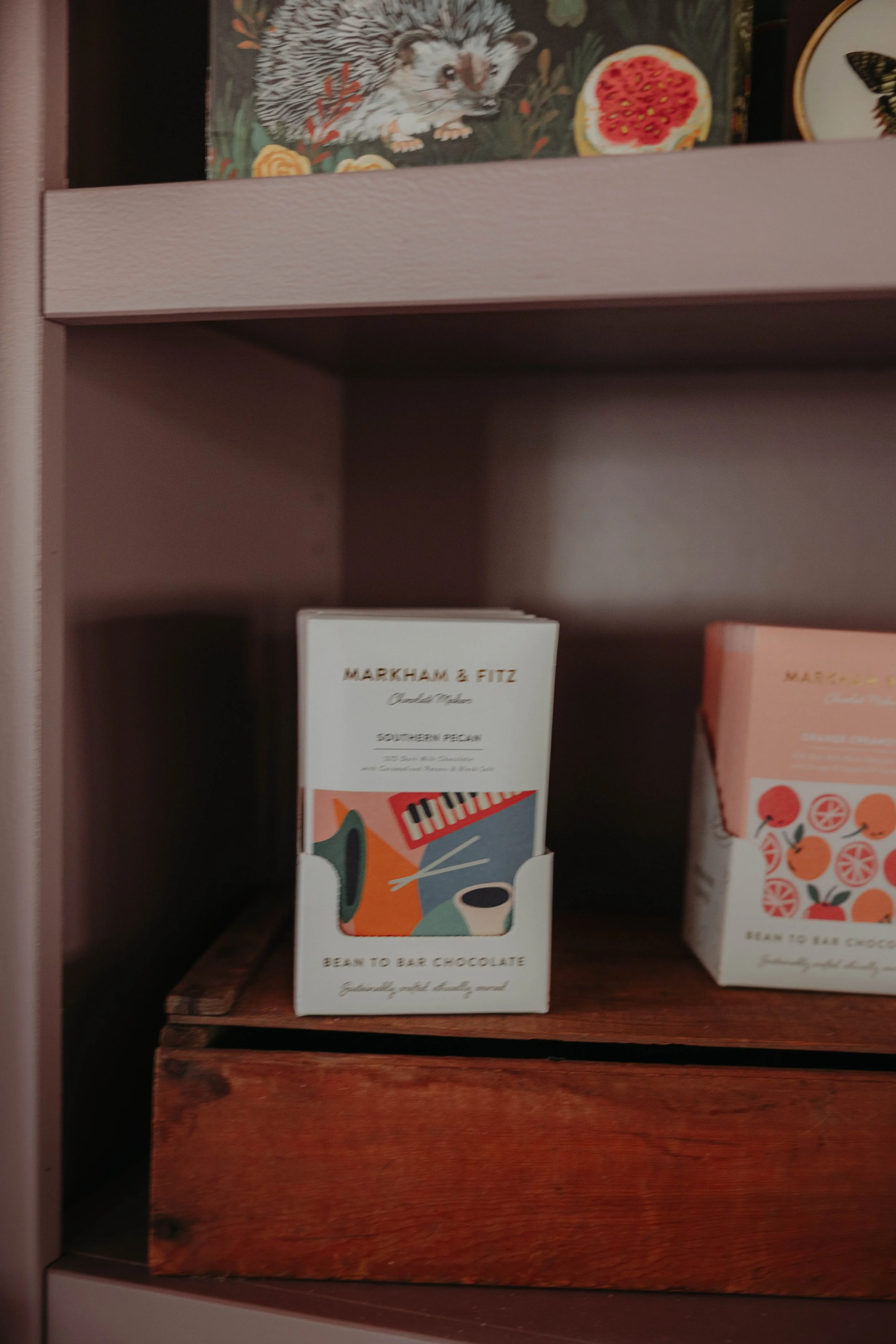Books on a wooden shelf, including a white book titled 'Marham & Fitz' with a colorful illustration of a musical instrument and a cup, and other books with partially visible titles and colorful patterns.