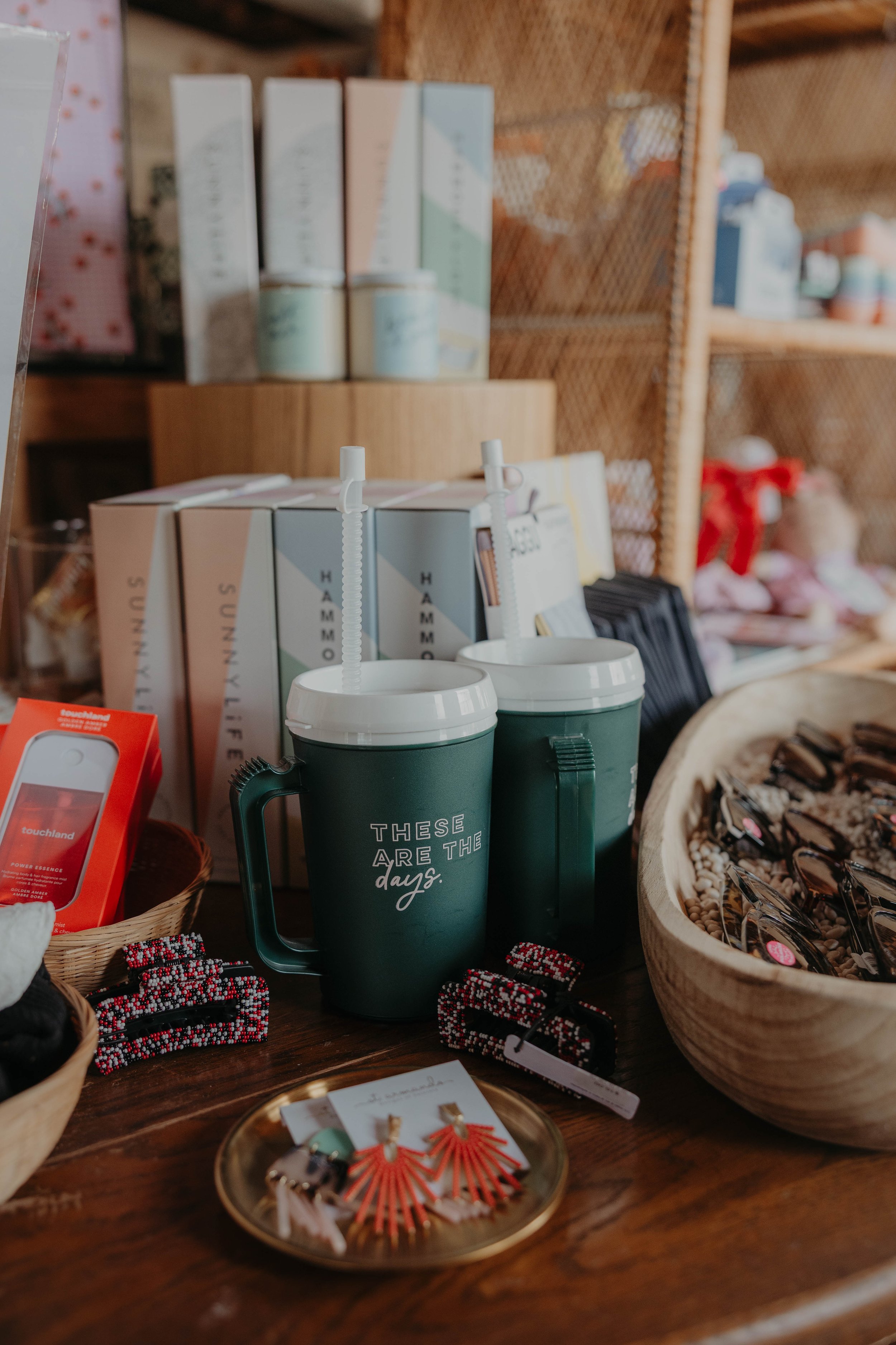 A display of items on a wooden table, including two green coffee mugs with straws, bracelets, jewelry, and various packaged products in the background, with shelves holding more gift items.