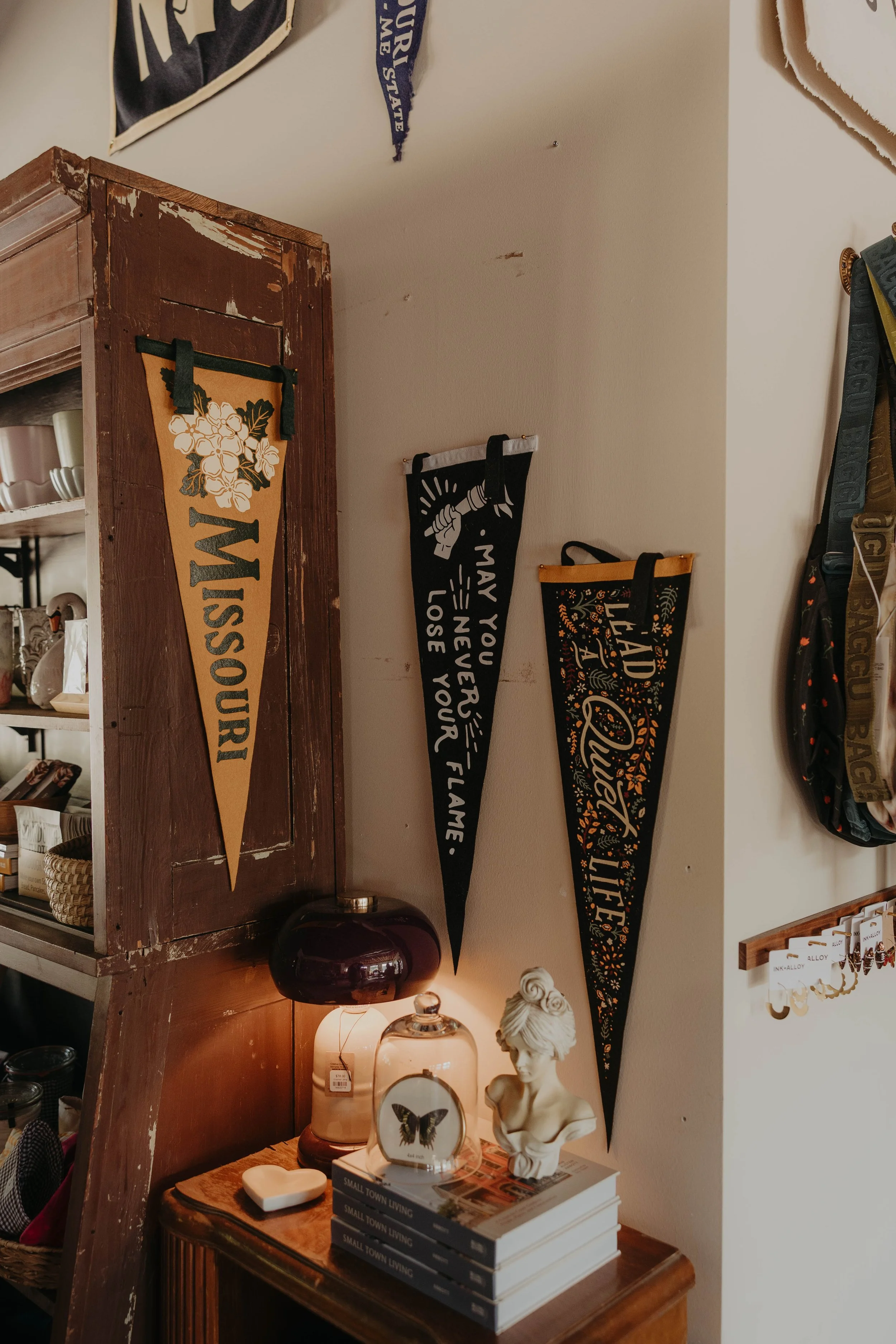 Decorative items in a home decor setting, featuring hanging banners with text, a wooden shelf with a vintage finish, a bust sculpture of a woman, a butterfly picture on a glass orb, lamps, and stacked books titled 'Small Town Living'.