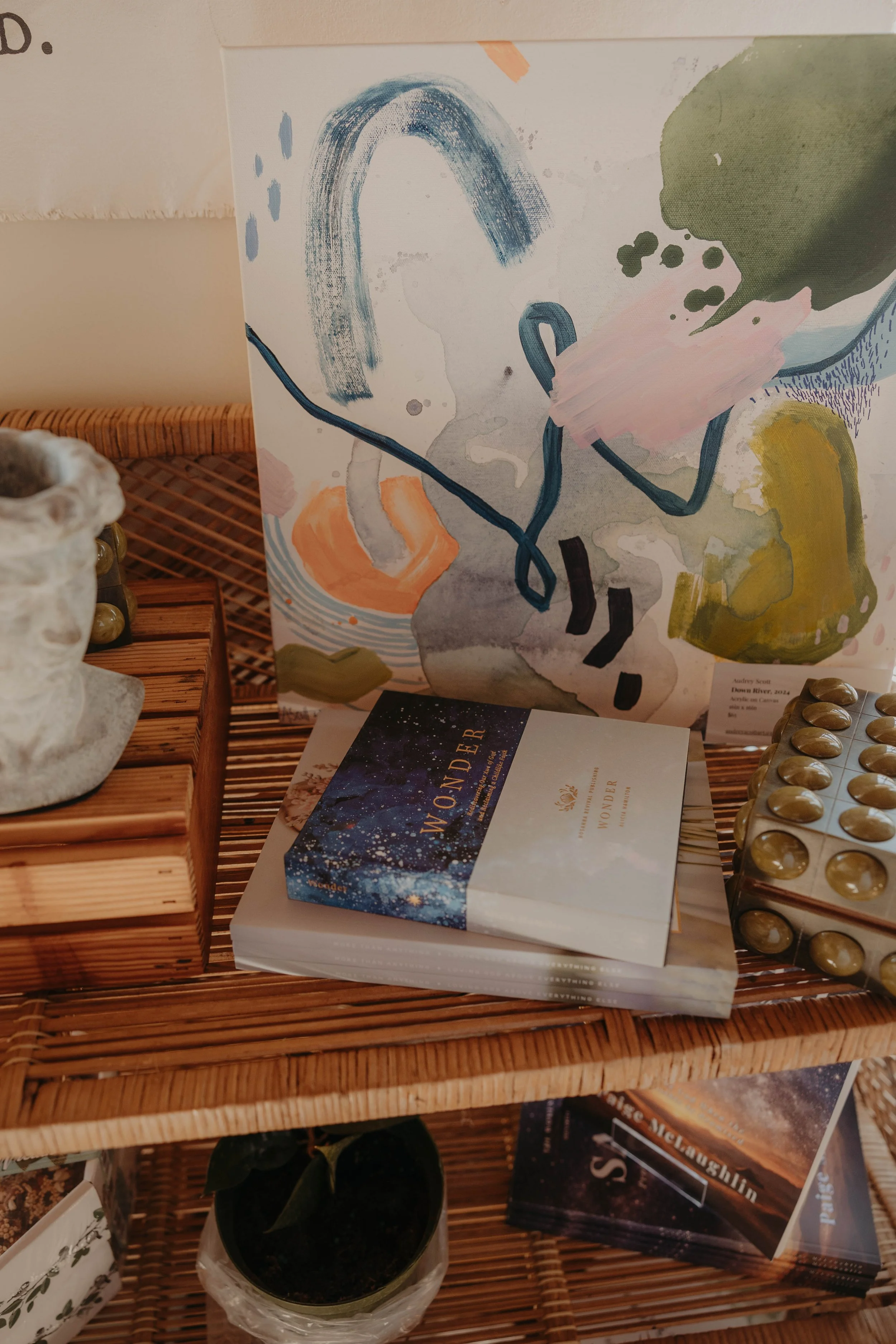 A colorful abstract painting with a rainbow, blobs, and brushstrokes displayed on a wicker shelf with books and decorative items.