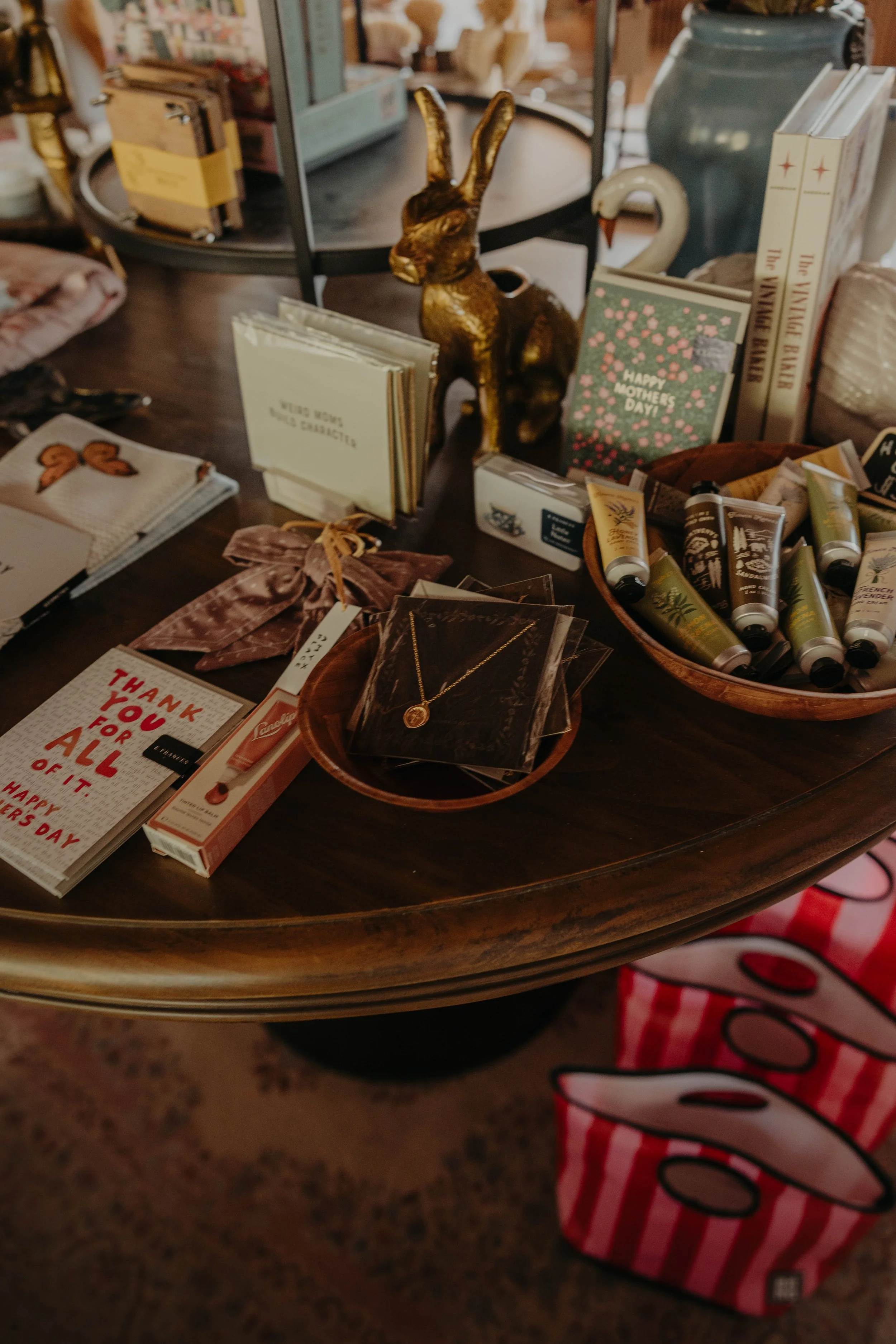 A table displays various greeting cards, a golden bunny figurine, skincare products, a necklace, and a thank you card. In the background, shelves with books and decorative items are visible, along with a swan-shaped figurine.