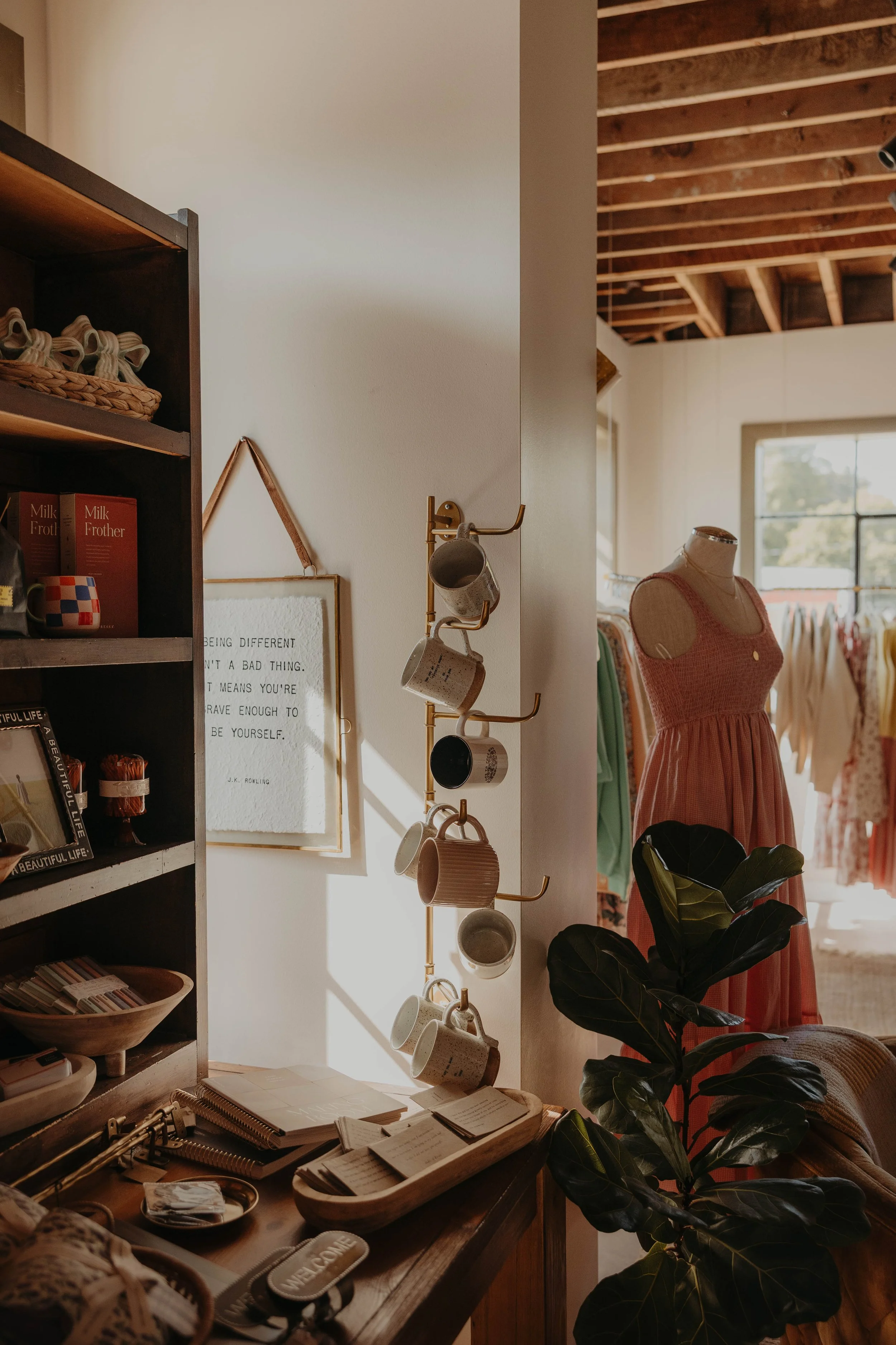 Interior of a boutique or home decor store with shelves, a mannequin wearing a pink dress, and various mugs hanging on a golden stand. Sunlight fills the room through a large window.