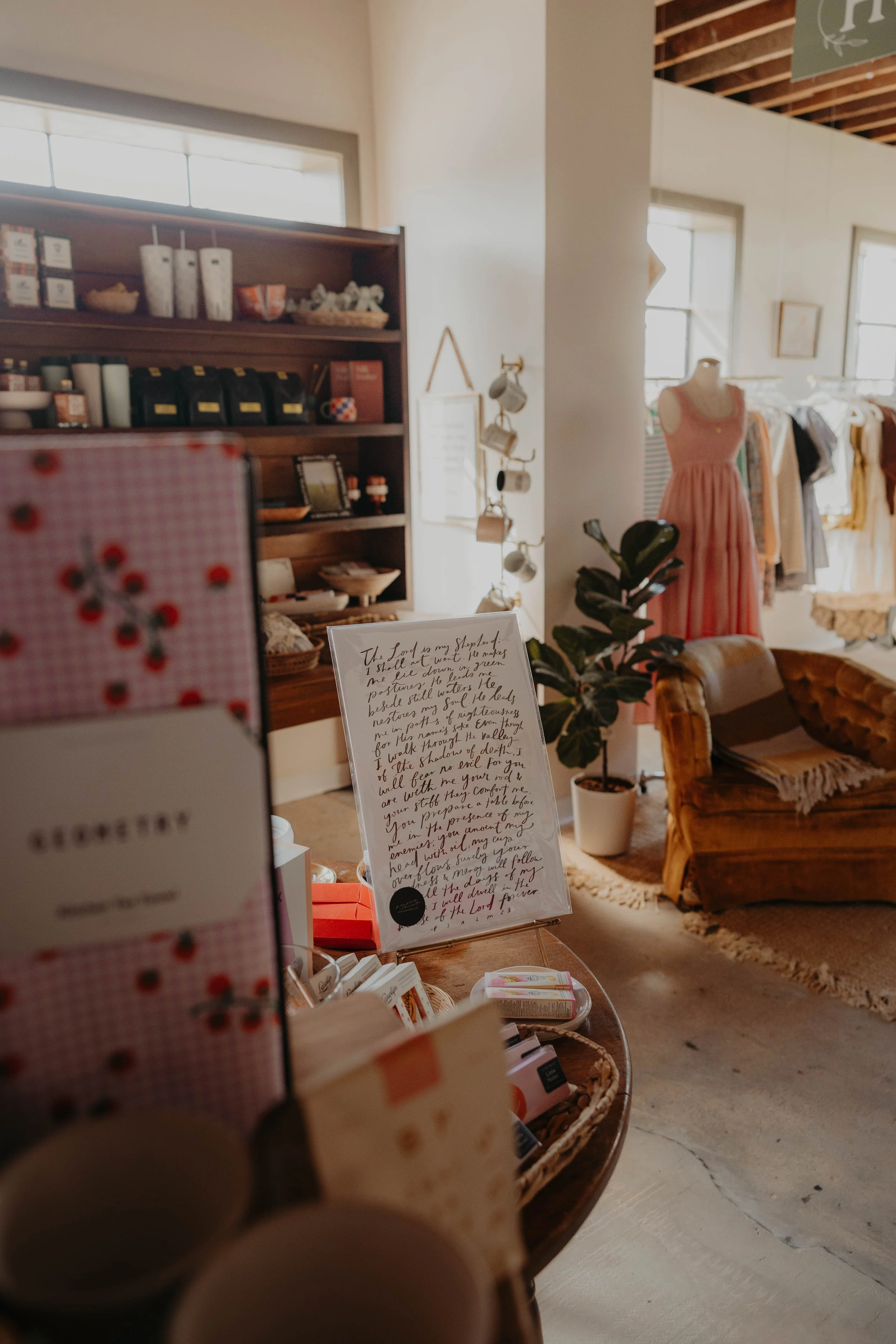 Inside a boutique store with wooden shelves, a mannequin dressed in a peach-colored dress, a cozy armchair, and various retail items including books, clothing, and decorative objects. There are windows allowing natural light to illuminate the space, 