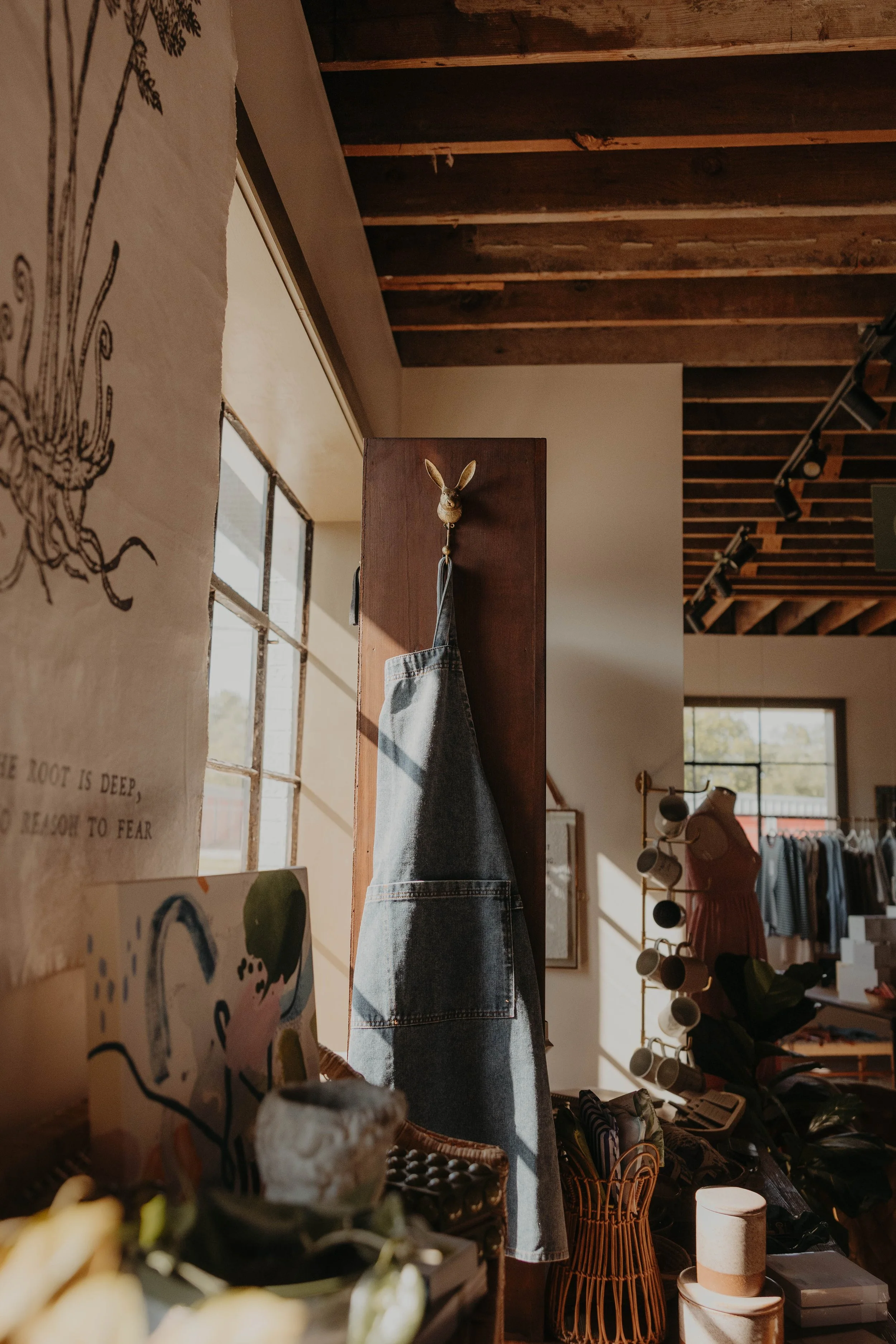 Interior of a retail store with wooden ceiling beams, natural light coming through large windows, and various items on display including a denim jacket hanging on a wooden post topped with a rabbit head ornament, and decor such as cups, baskets, and 