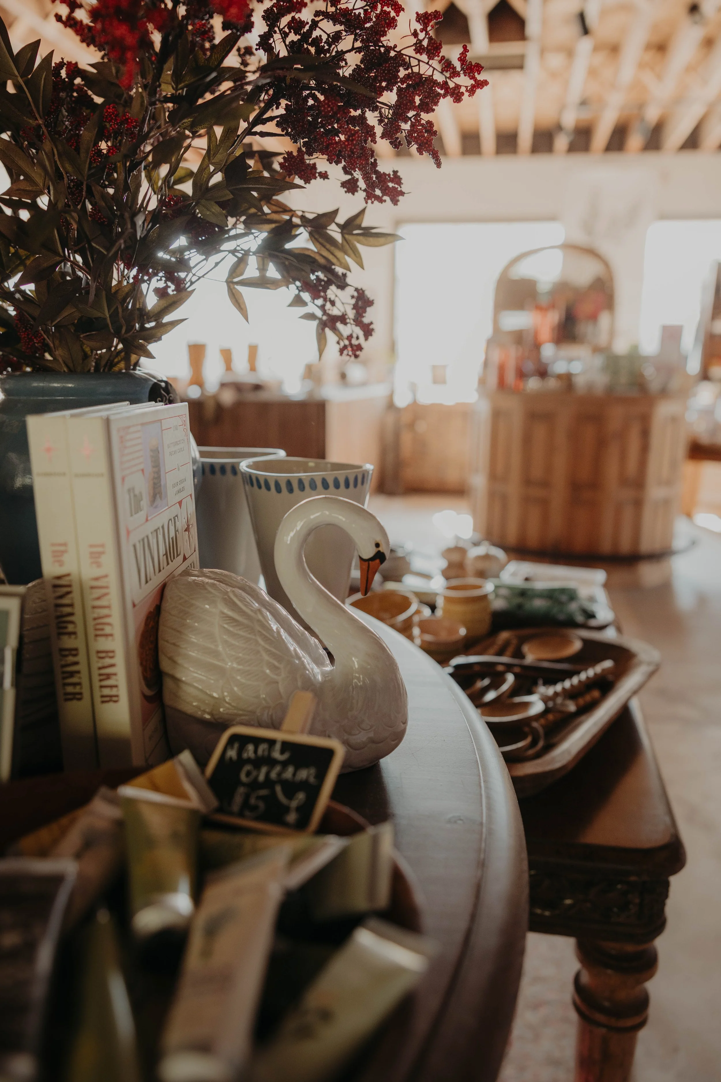 Display of various home decor items and kitchenware on a wooden table in a cozy, well-lit shop. Items include a ceramic swan, books, and candlesticks, with blurred furniture and shelves in the background.
