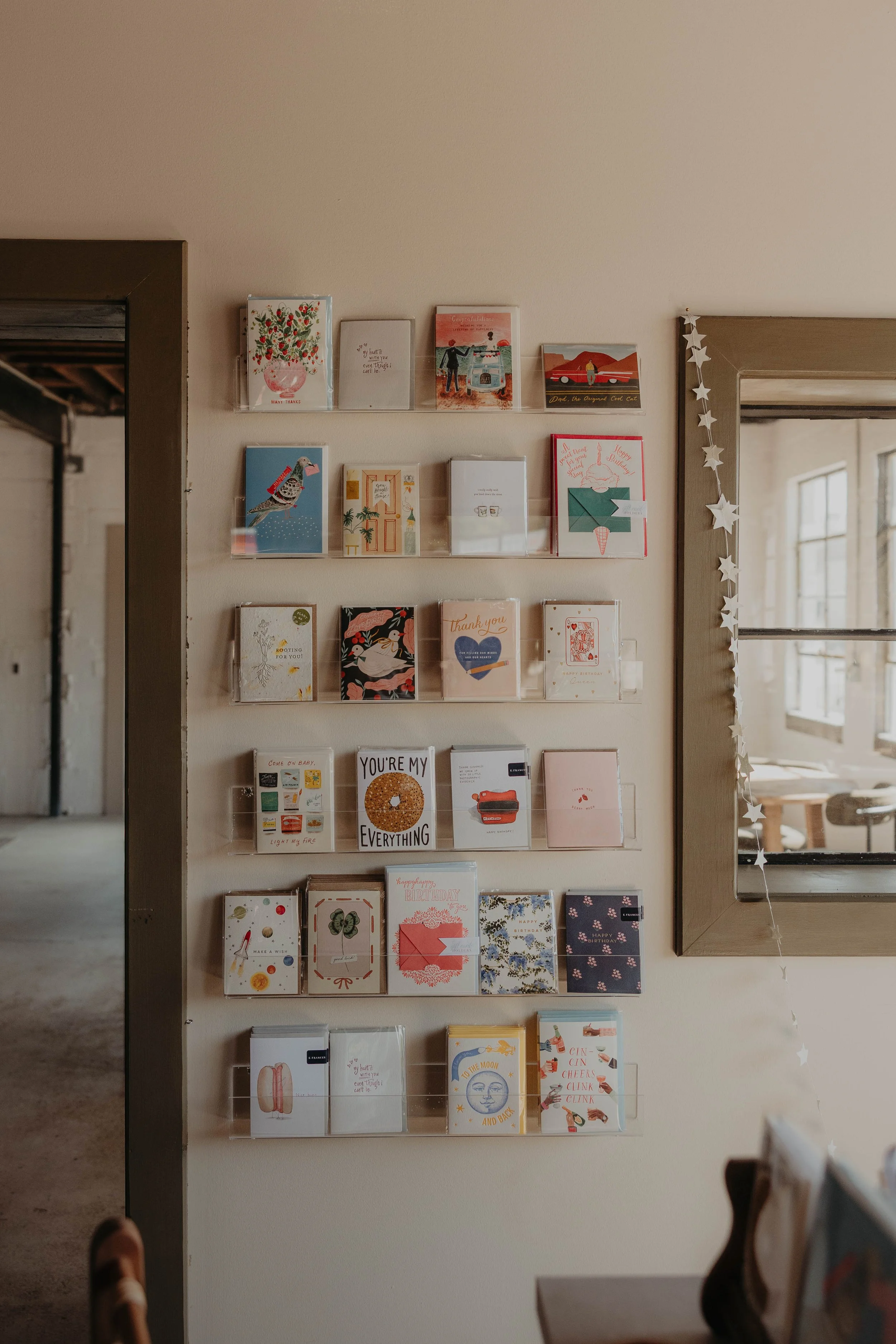 A display of greeting cards on a wall, arranged on clear plastic shelves, next to a mirror with star-shaped garland. The cards feature various illustrations and messages, likely for occasions like birthdays and holidays.