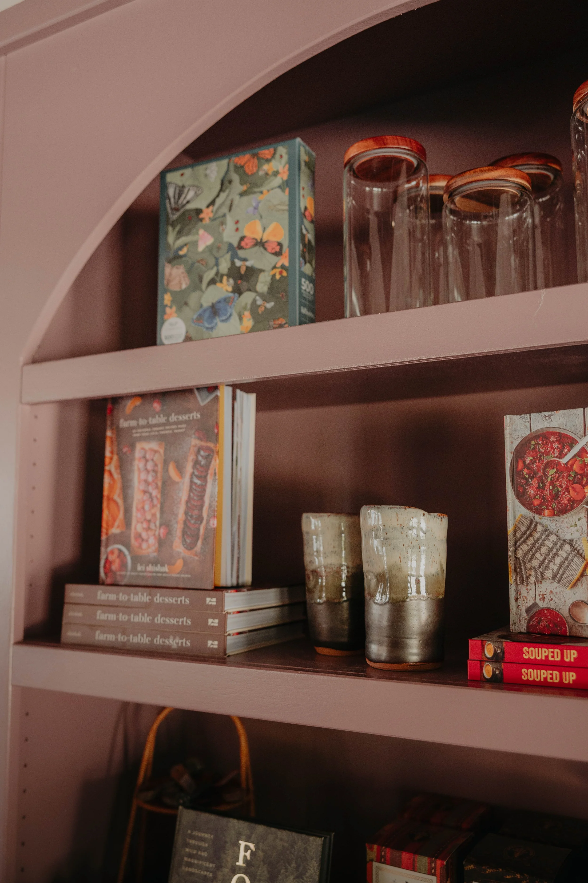 A pink bookshelf containing books, three ceramic cups with a rustic glaze, and clear glasses with wooden lids.