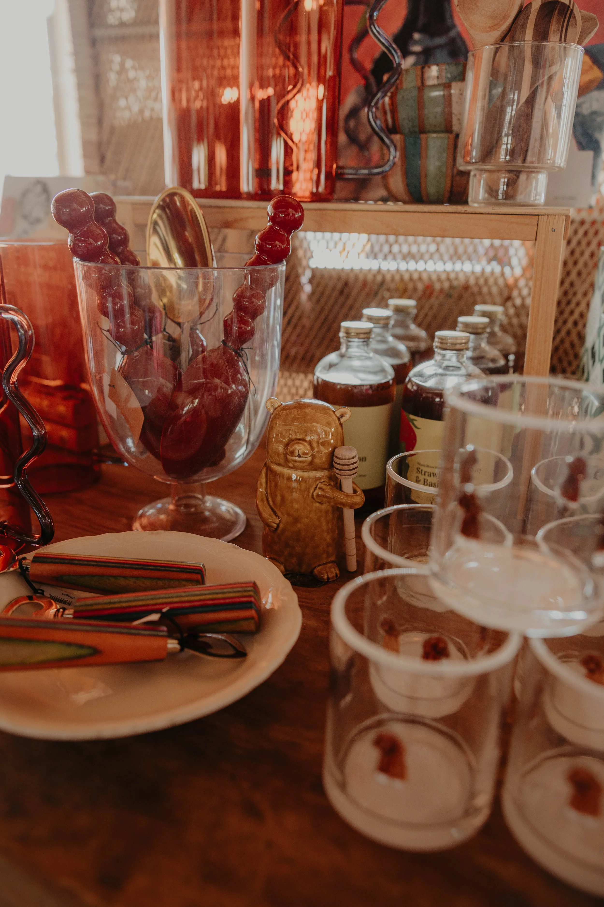 A display of various items on a wooden table, including glasses filled with red candies, small bottles of drinks, a ceramic bear figurine, and colorful striped napkins, with a wooden shelf behind holding glass jars and containers.