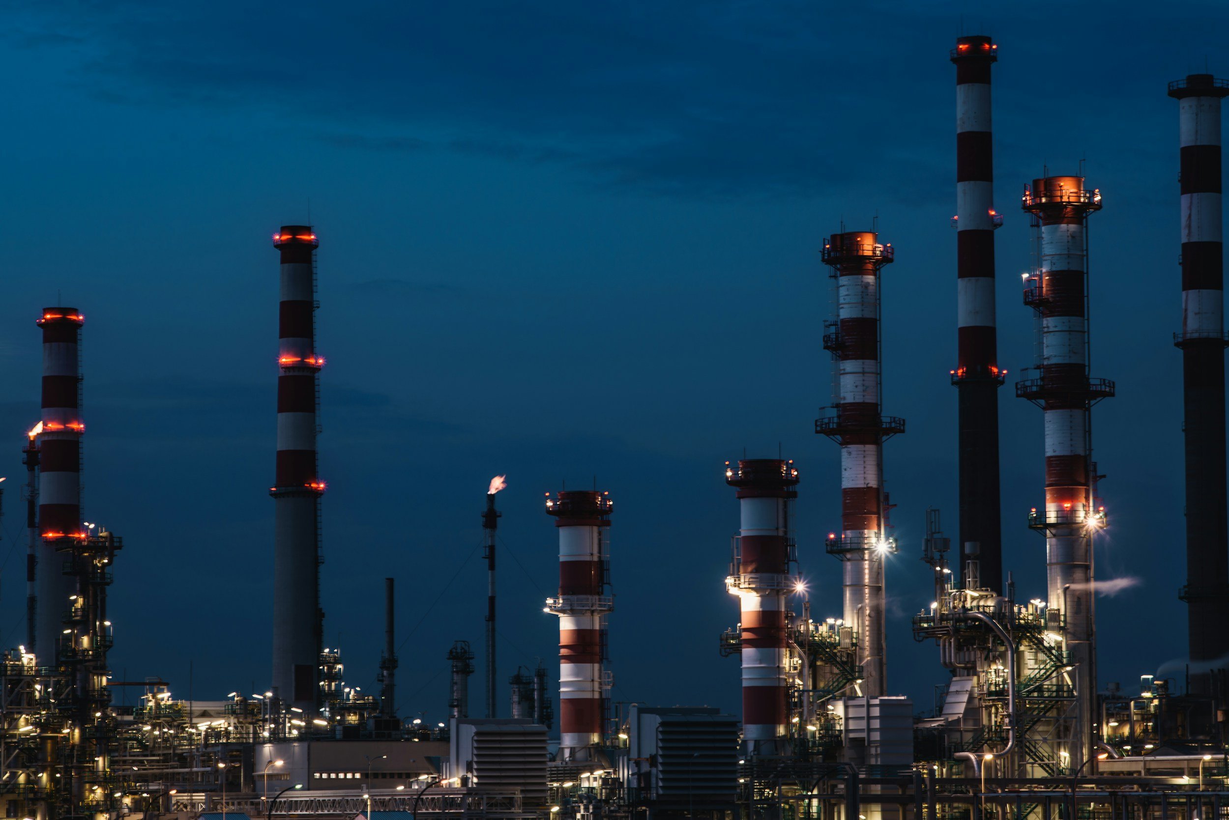 Nighttime view of an industrial oil refinery with tall smokestacks illuminated against a dark sky.