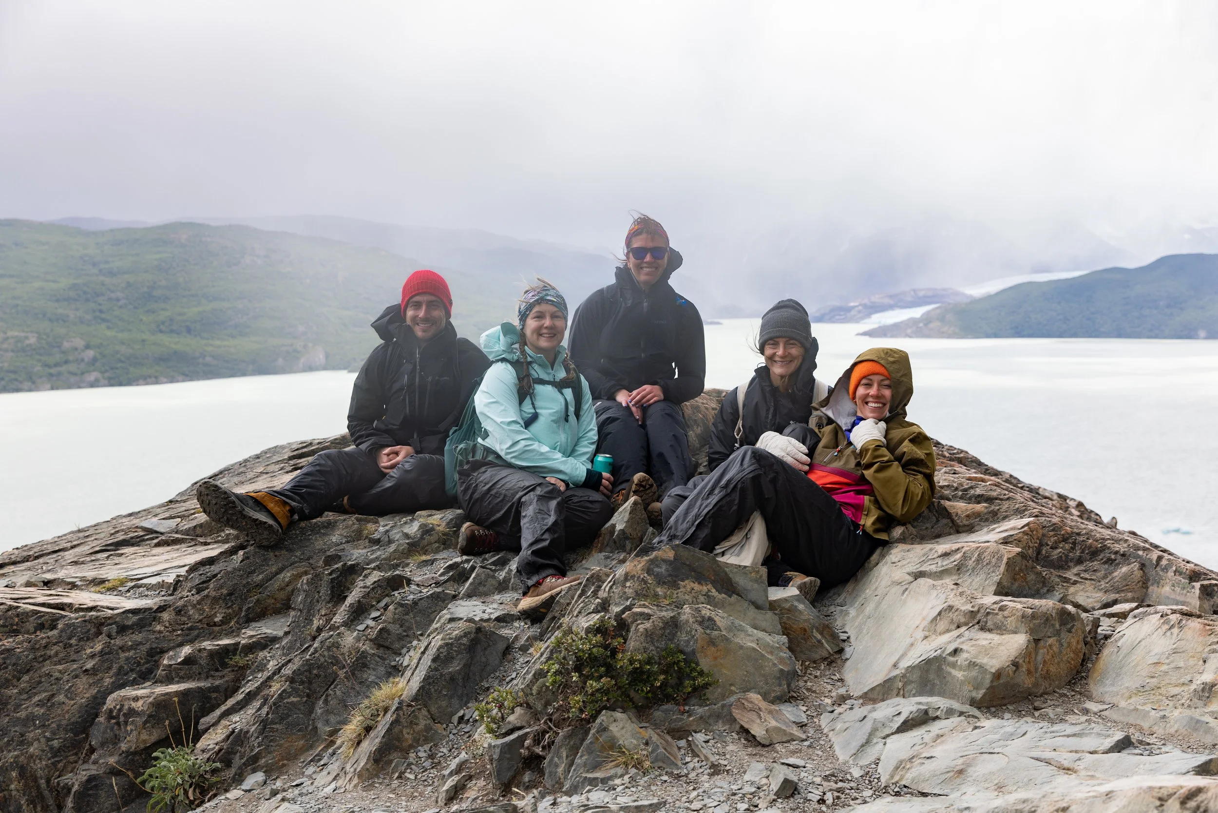 Group of five hikers in outdoor gear sitting on rocks near a mountain lake with a cloudy sky.