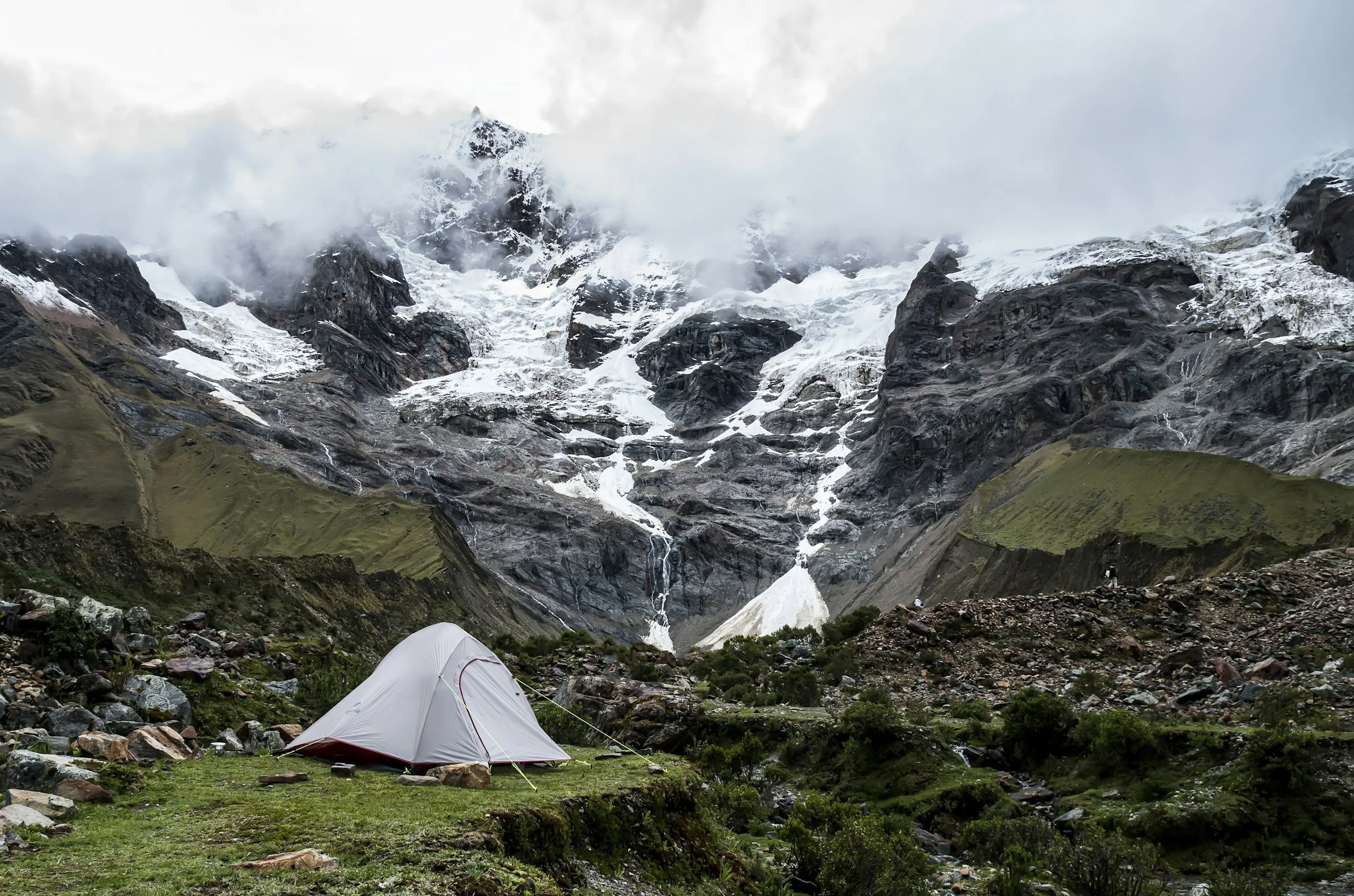 A tent set up on grassy land at the base of snow-capped mountains, with a waterfall visible in the distance and clouds covering the peaks.