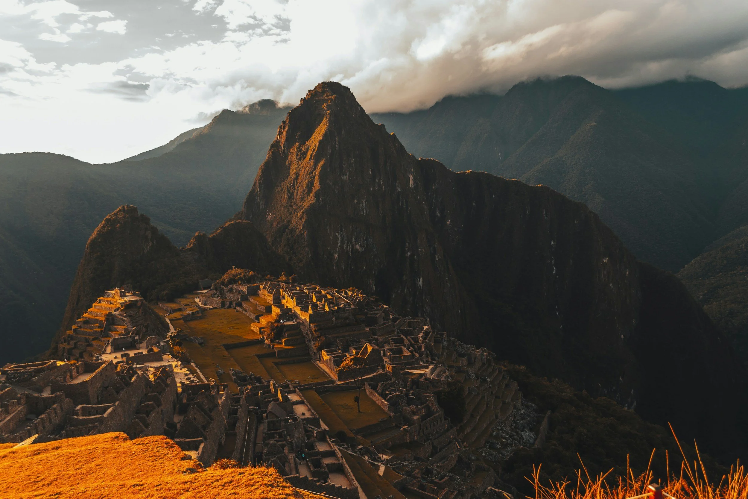 Sunset over Machu Picchu, with the ancient Incan ruins on a mountain ridge surrounded by lush greenery and misty mountains in the background.