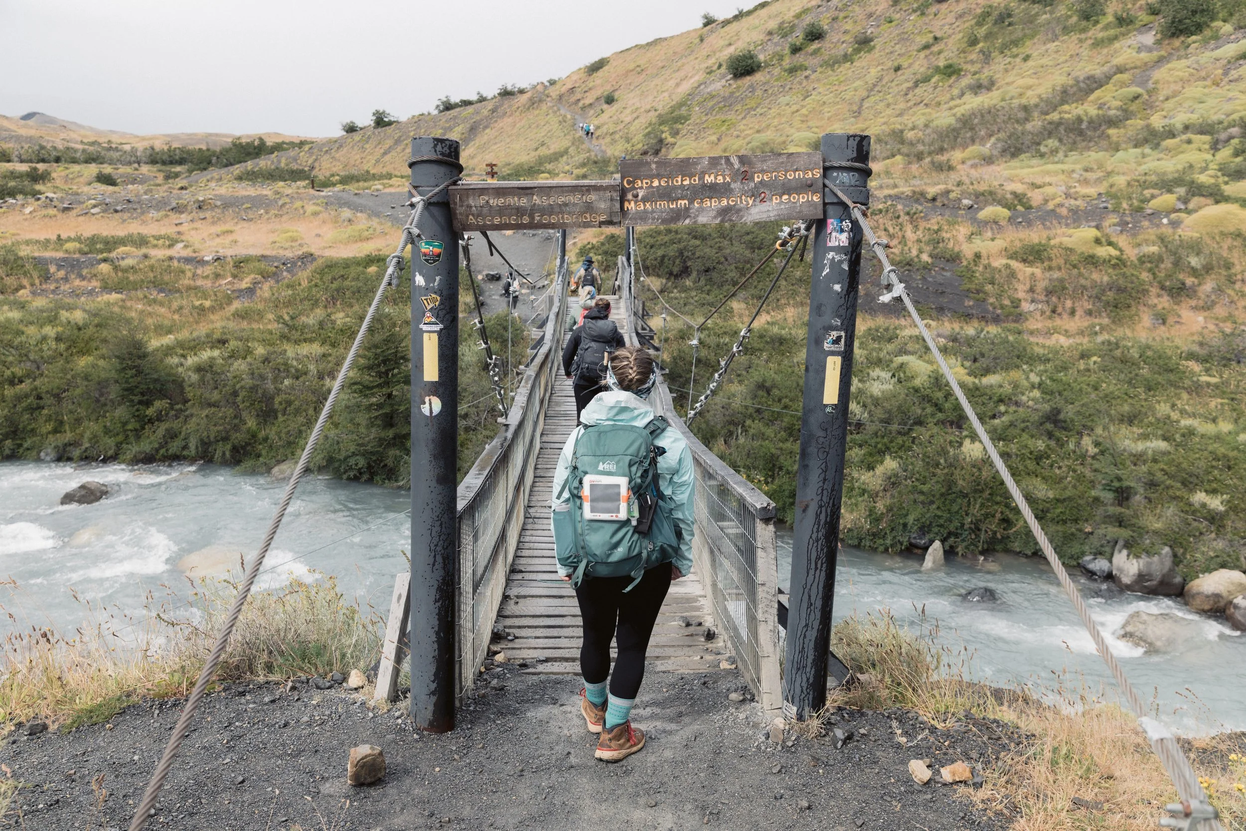 Hikers walking across a suspension footbridge over a river in a mountainous area.