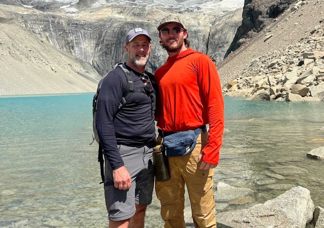 Two men standing in a clear blue lake surrounded by rocky mountains, smiling at the camera. One wears a black long-sleeve shirt and gray shorts, the other a red long-sleeve shirt and tan pants.