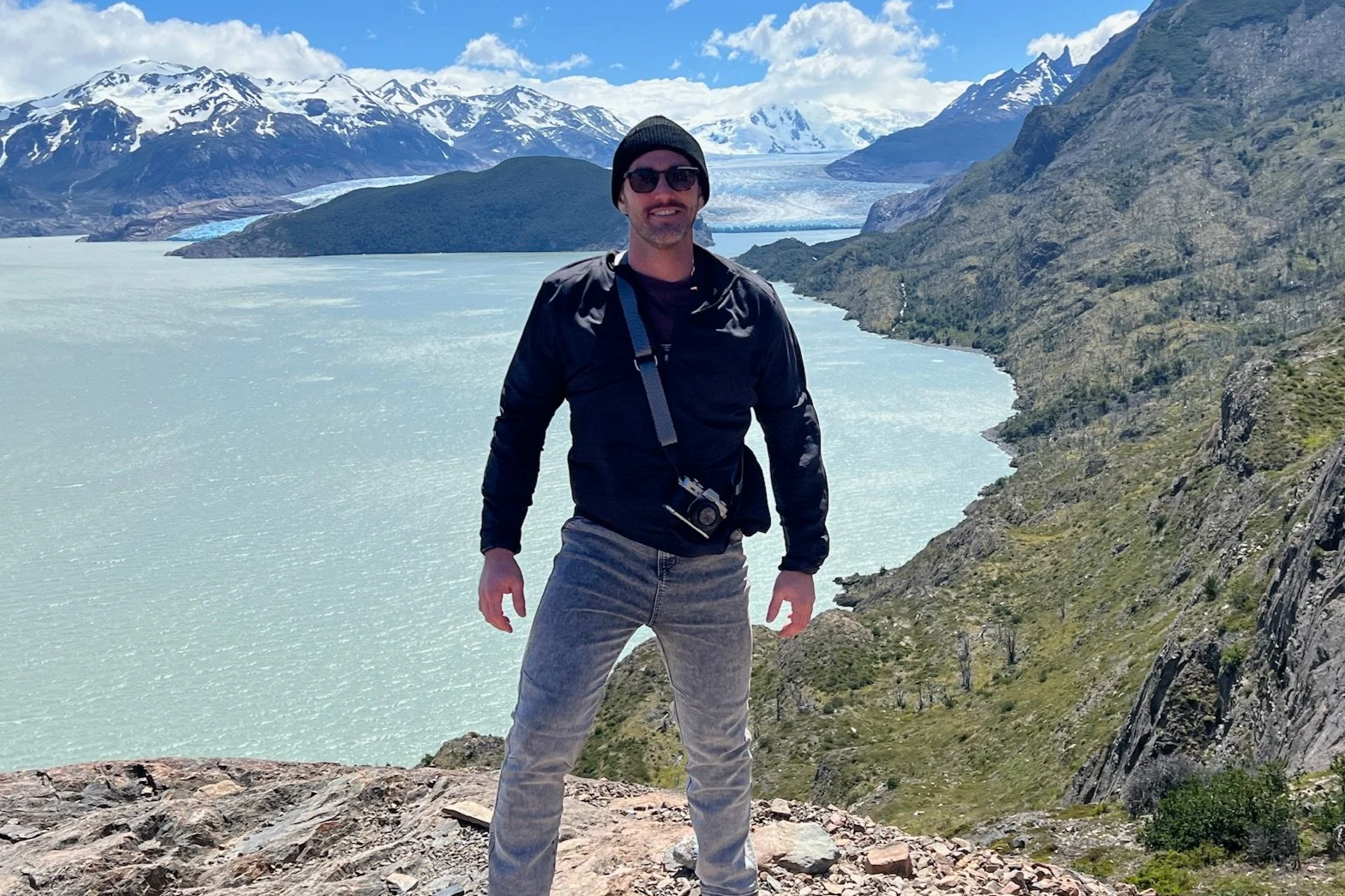 A man wearing sunglasses, a black beanie, a black jacket, and gray jeans standing on rocky terrain with snow-capped mountains, a glacier, and a body of water in the background.