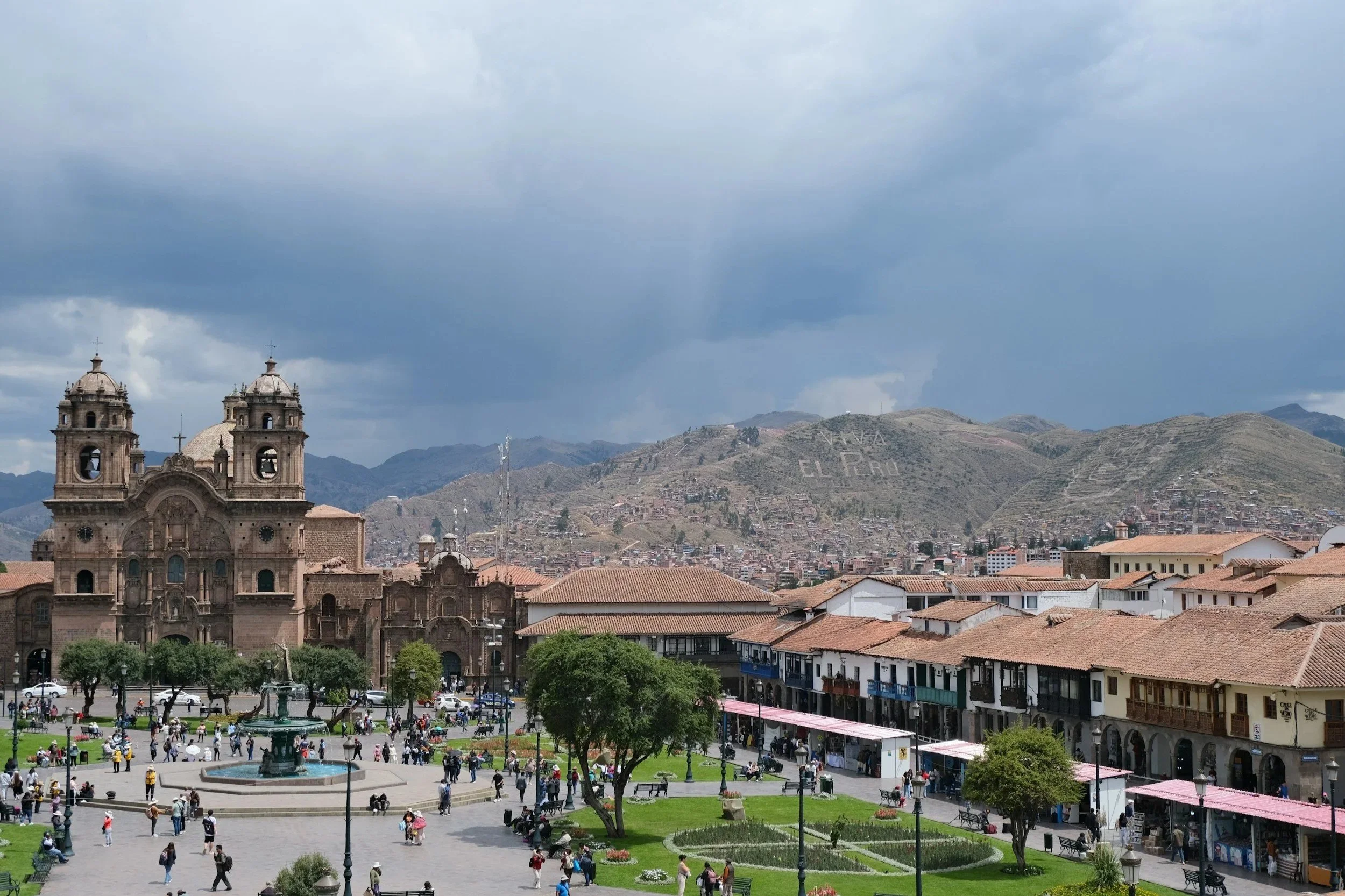 City square with a historic church, green park with trees and a fountain, and colorful buildings with shops, set against a mountainous background with cloudy sky.