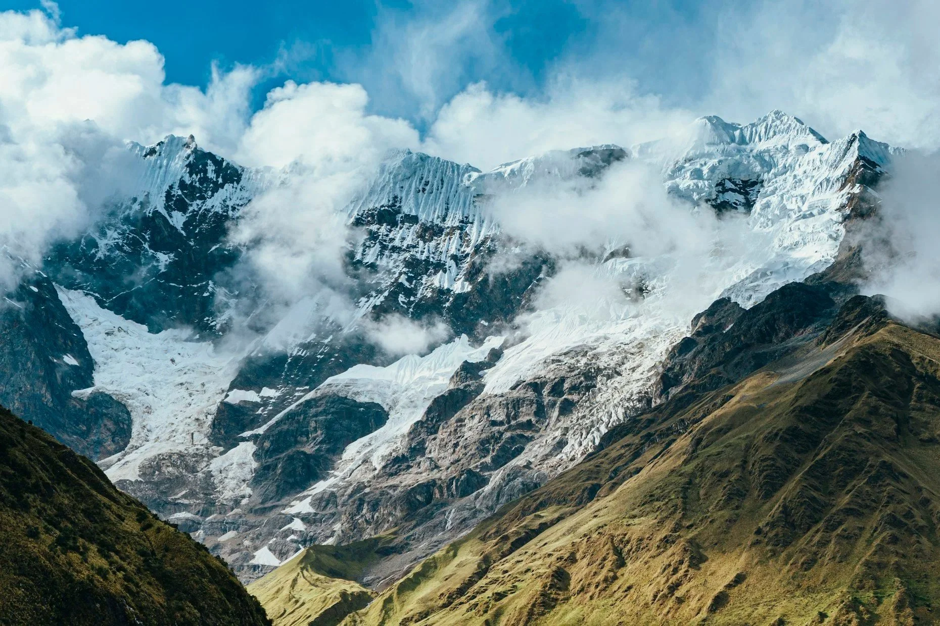 Snow-covered mountain peaks with clouds in the sky and green grassy slopes in the foreground.