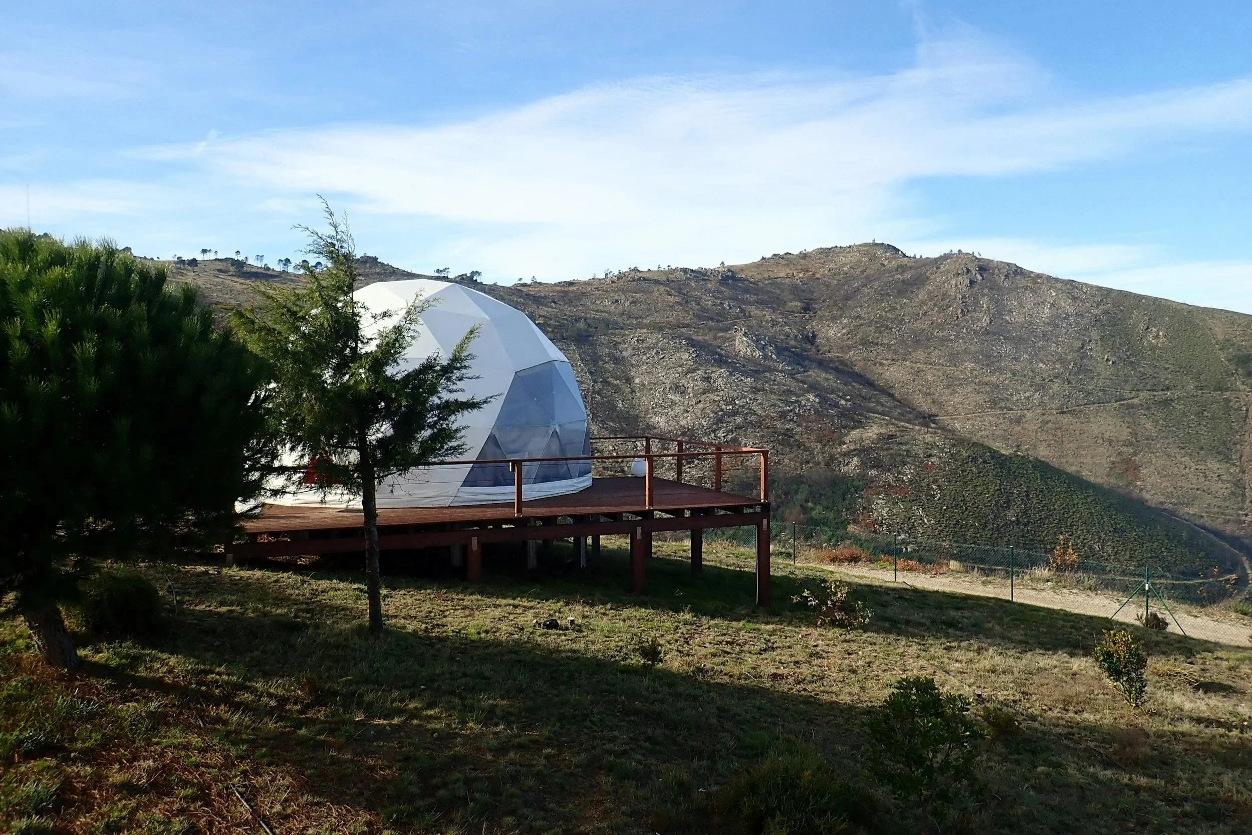 A geodesic dome structure on a wooden deck, situated on a grassy hillside with trees, against a mountain range under a cloudy sky.