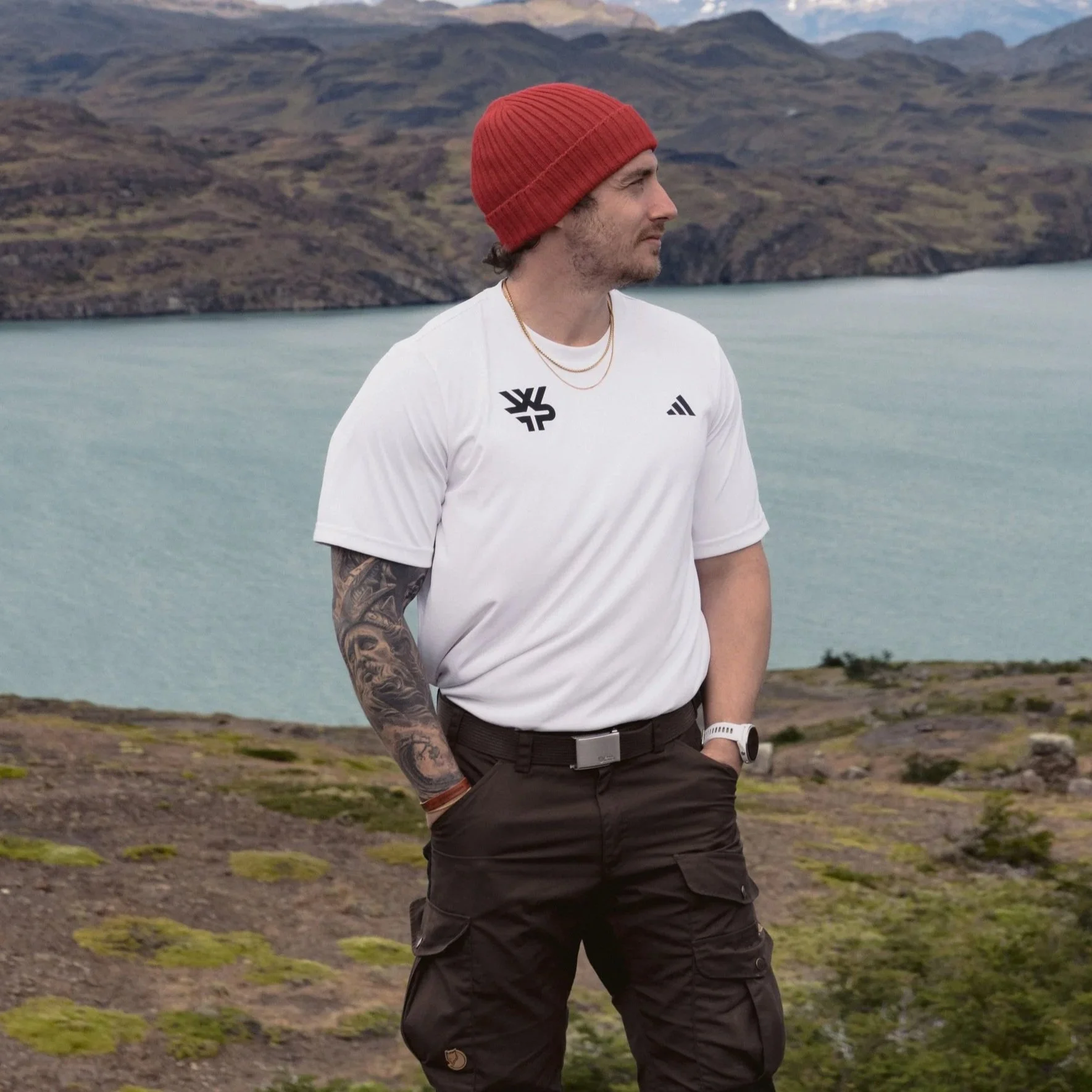 A young man standing outdoors with a lake and mountains in the background, wearing a red beanie, white t-shirt, cargo pants, and a watch, looking to the right.