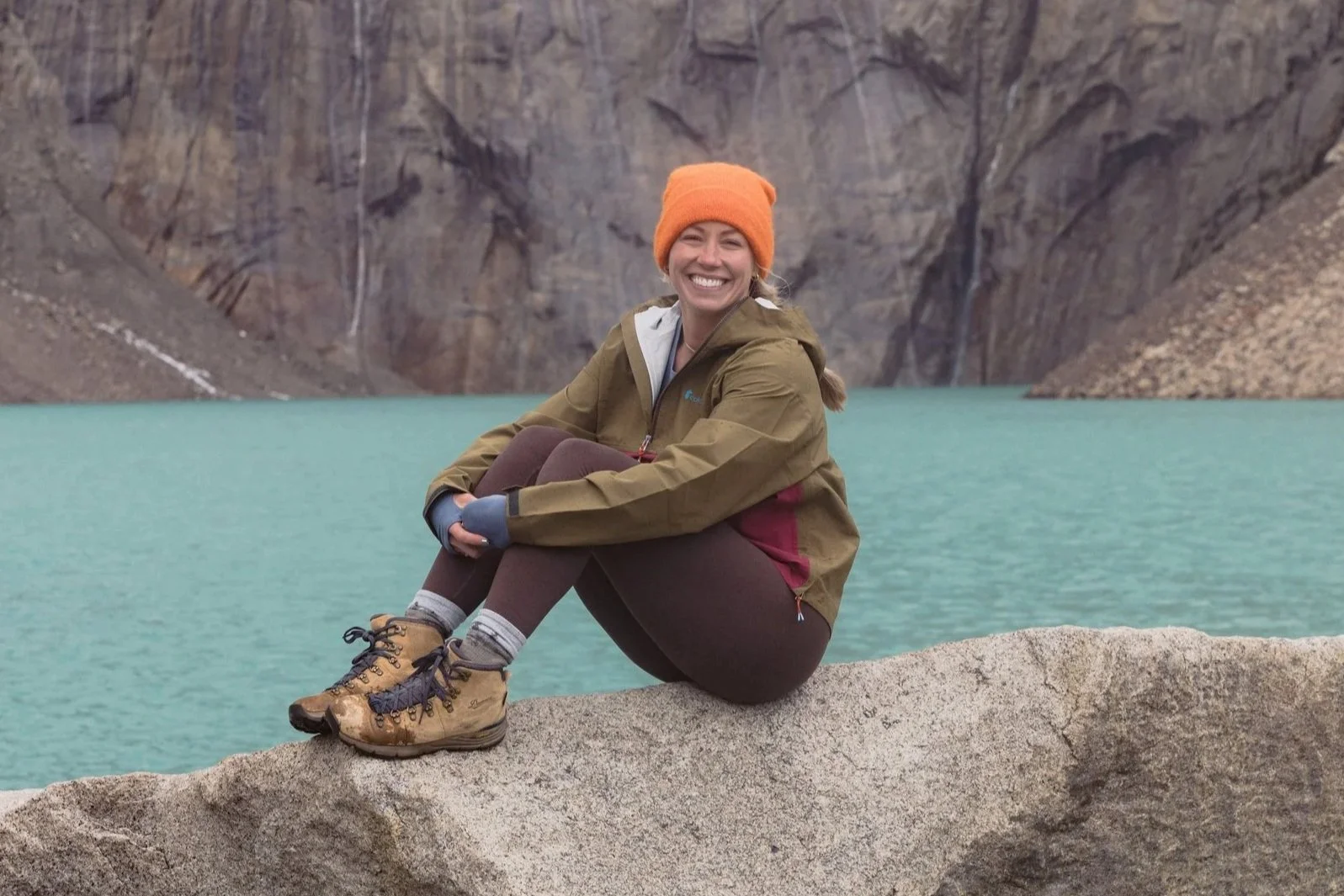 Smiling woman in outdoor gear sitting on a rock near a mountain lake with cliffs in the background.