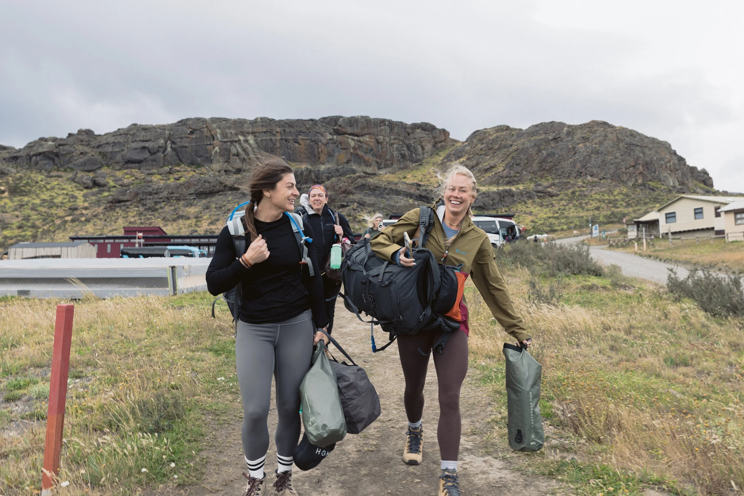Group of four women hiking outdoors on a trail, carrying backpacks and gear, smiling and talking, with rocky hills and residential buildings in the background.
