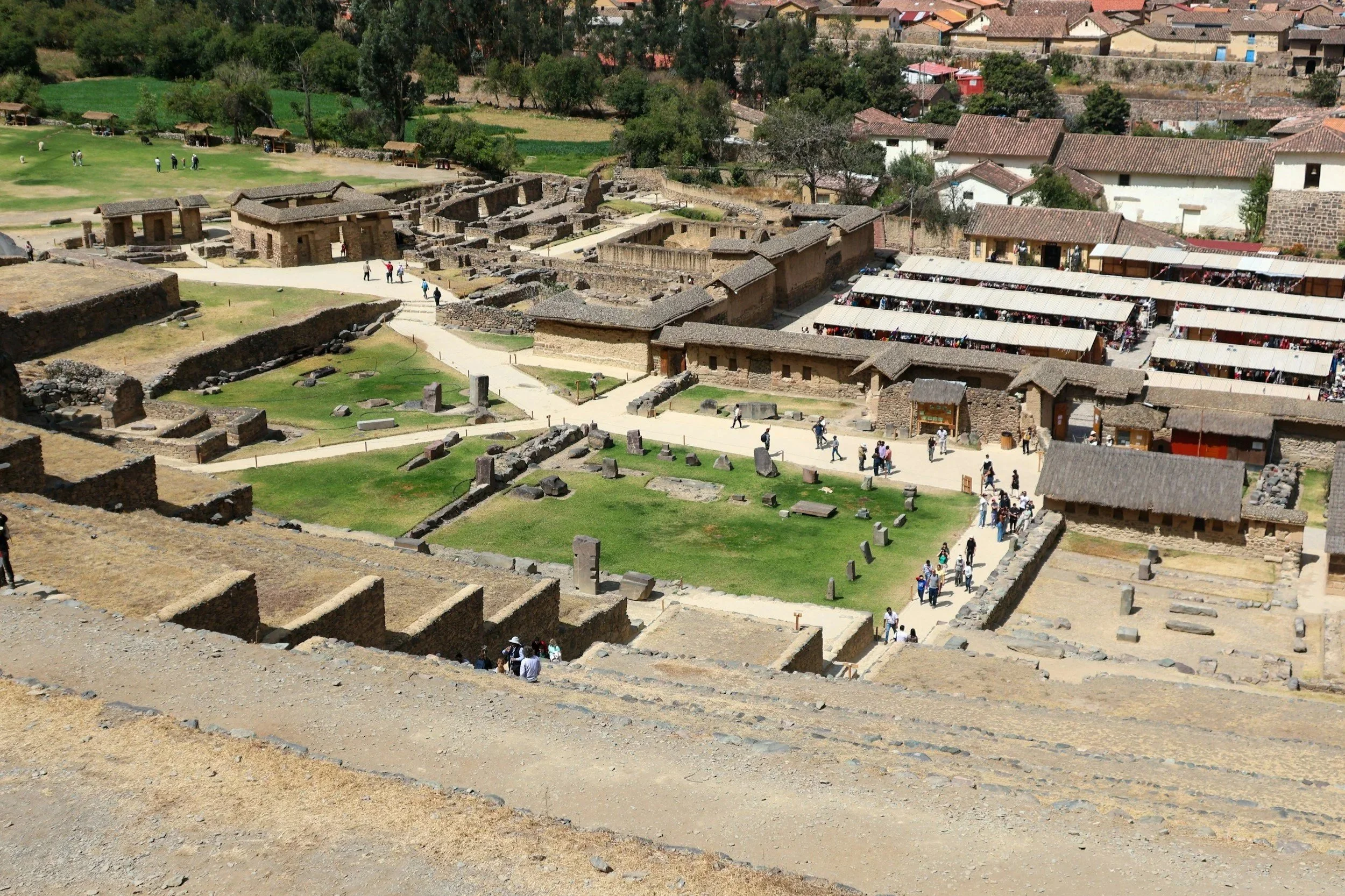 An archaeological site with ancient stone ruins, pathways, and grassy areas, with tourists walking and exploring the site, and buildings with thatched roofs nearby.